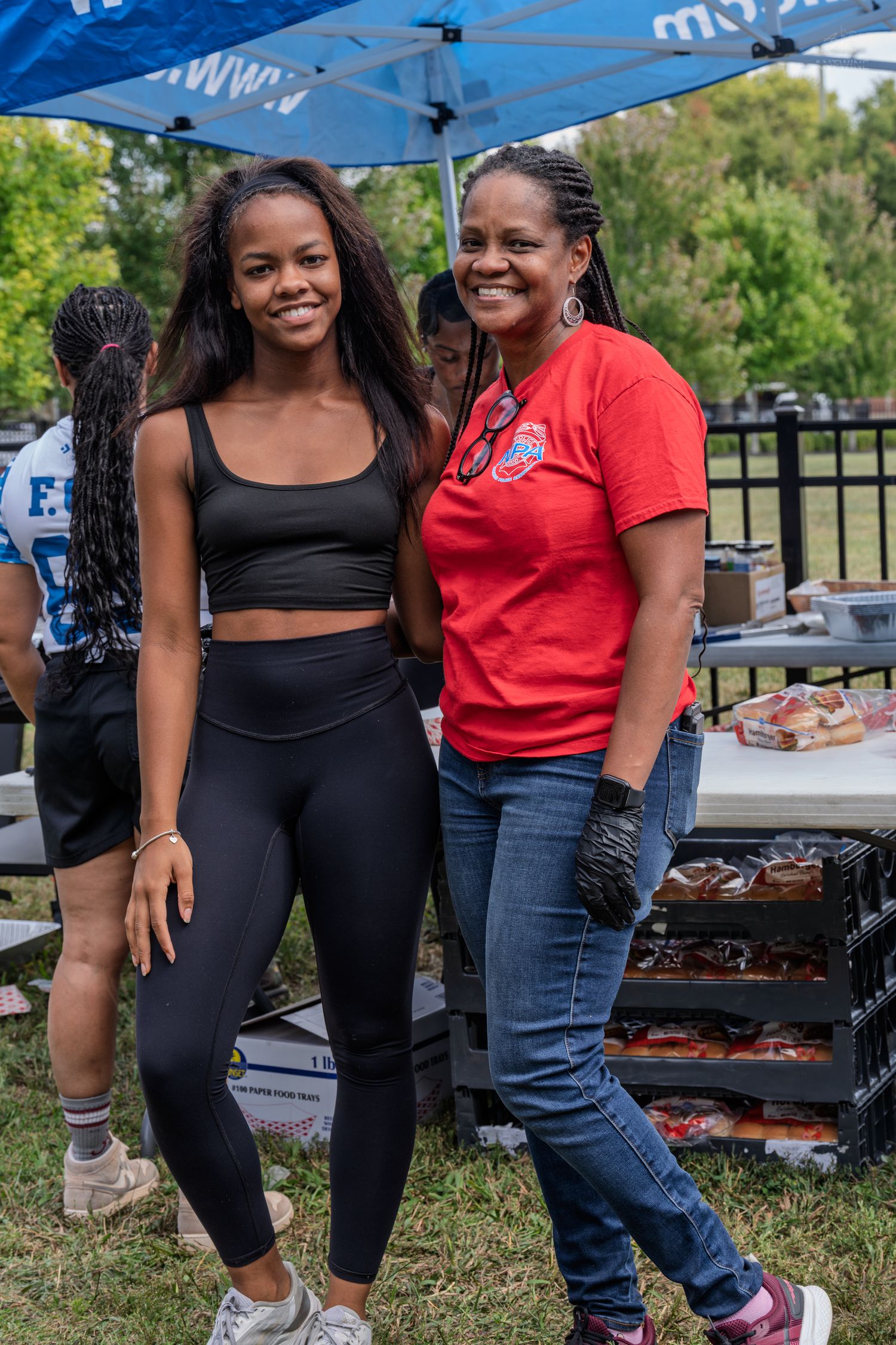 Two women pose together smiling at an outdoor community barbecue event under a blue canopy.