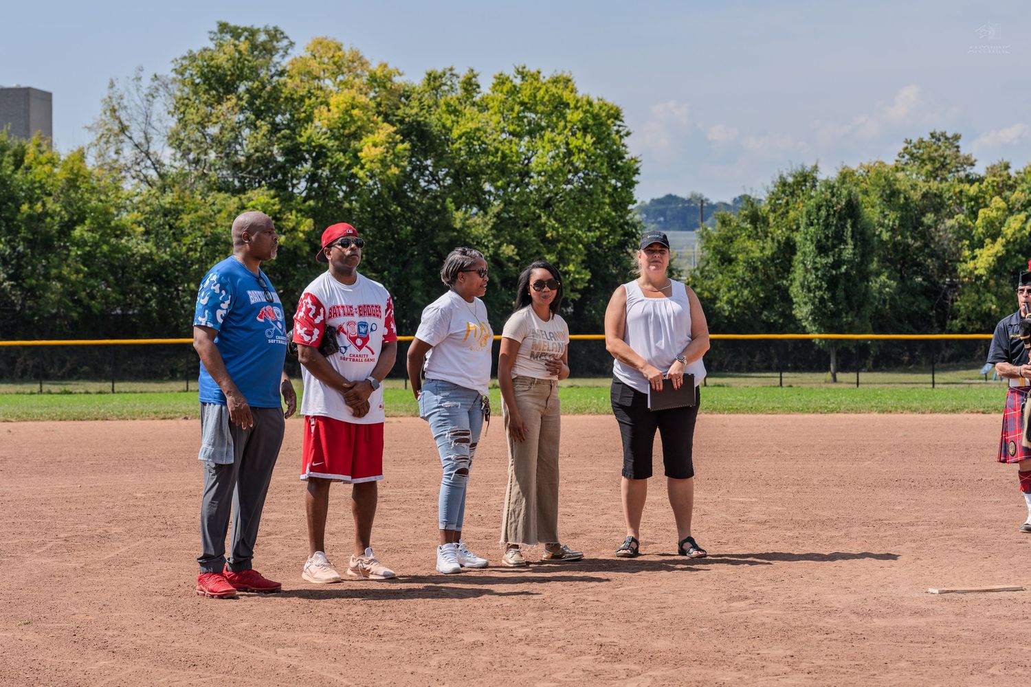 Five people stand together on a baseball diamond during an outdoor softball event on a sunny day.