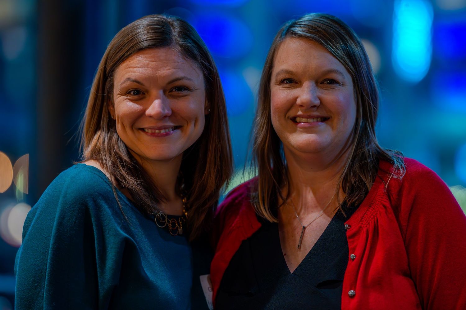 Two smiling women pose together at an indoor event, one in teal and one in red cardigan.