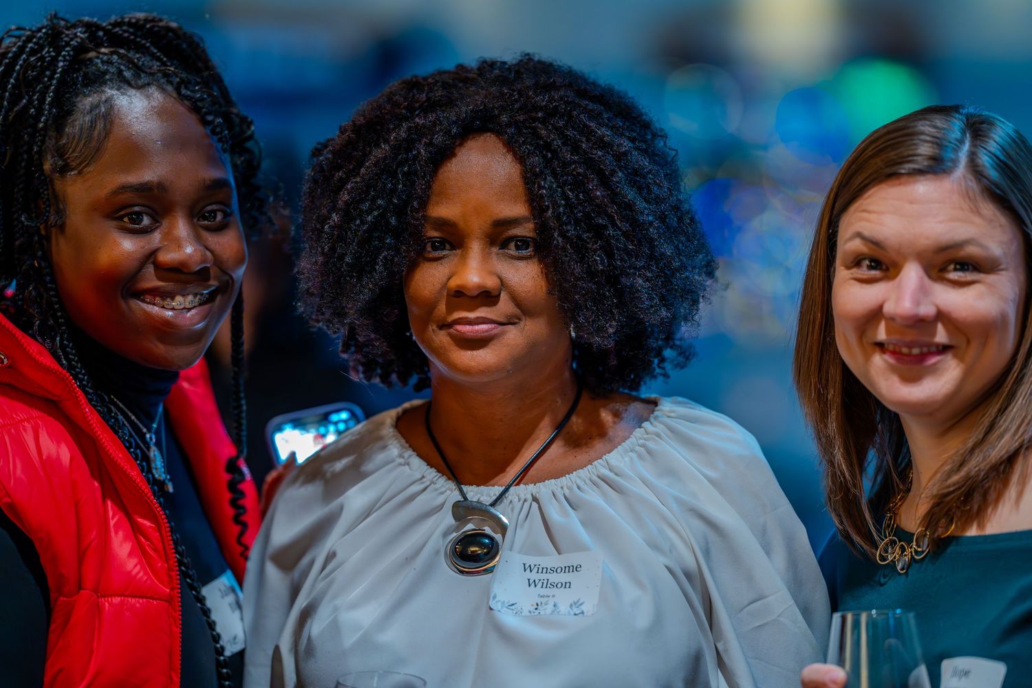 Three women smiling together at a networking event, posing for a photo in a warmly lit indoor venue.