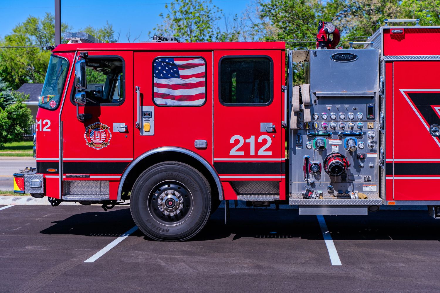 Red fire truck number 212 with American flag decal parked in lot, featuring equipment panels and hose connections on sunny day.