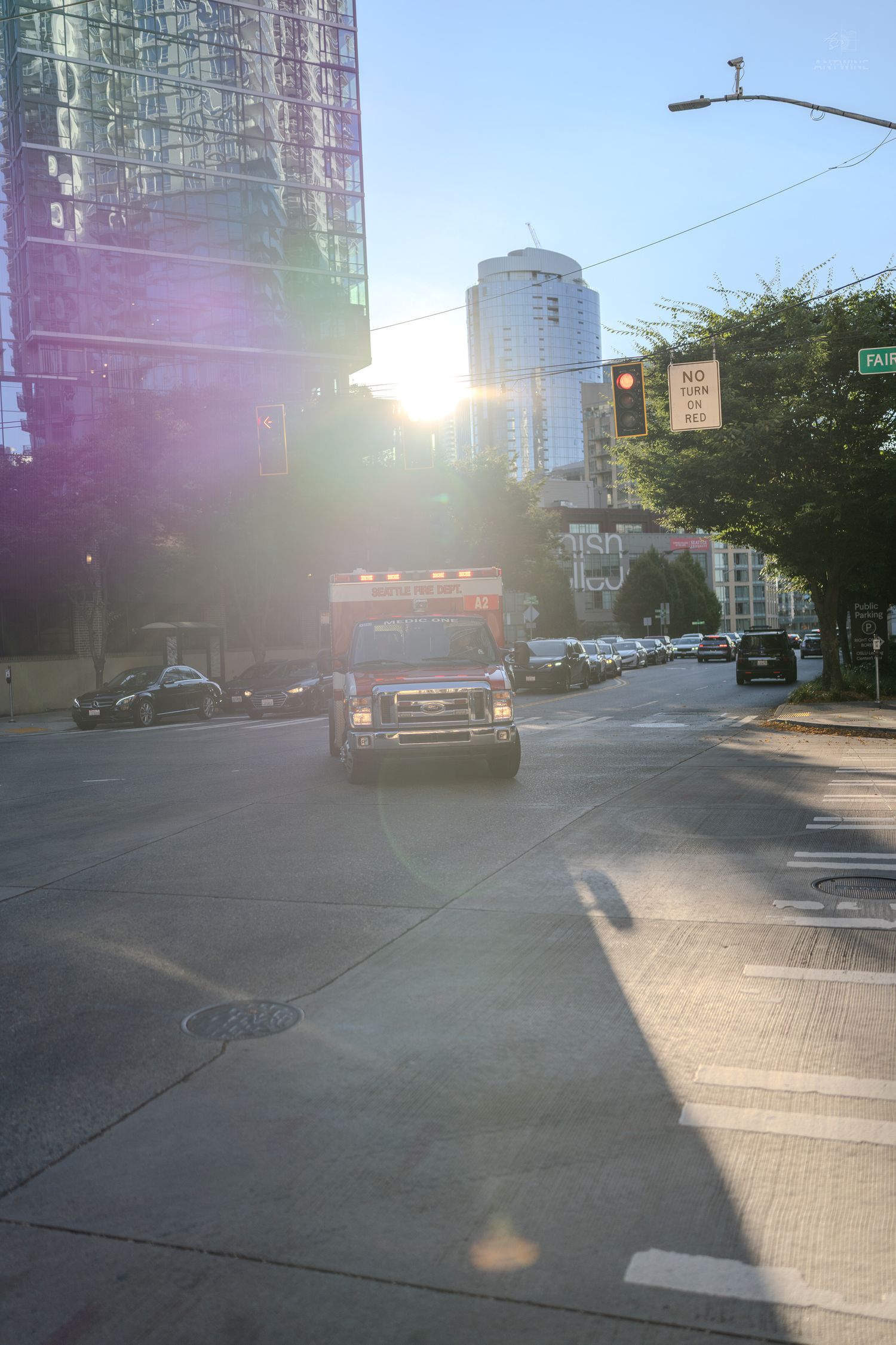 Emergency ambulance driving through sunny urban intersection with tall city buildings in background.