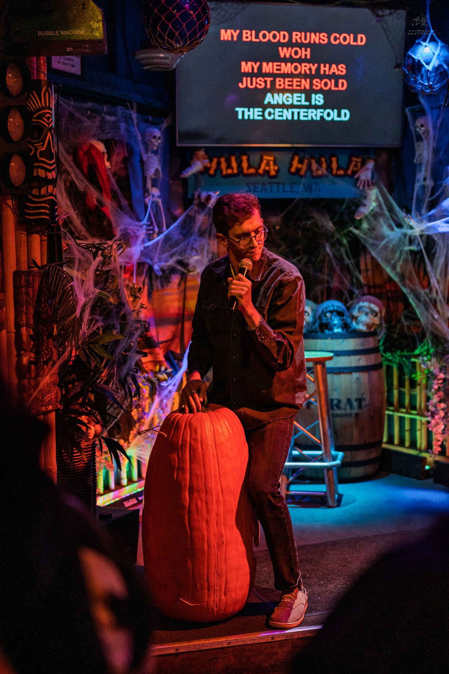 Man sings karaoke at Halloween bar with cobwebs, pumpkin, and lyrics screen showing Angel is the Centerfold.