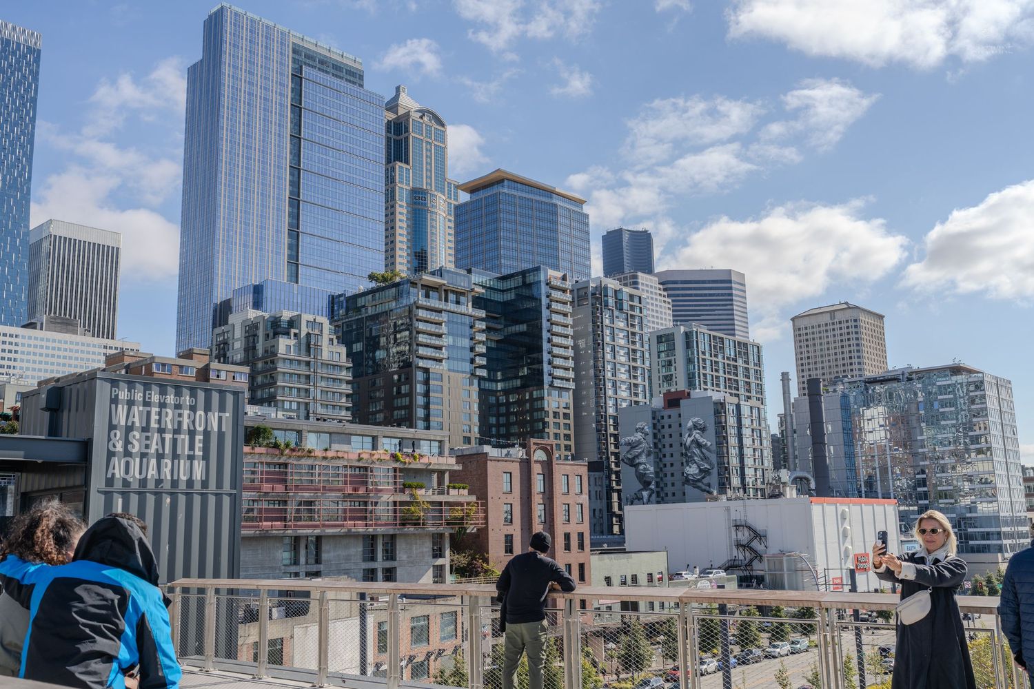 Tourists photographing Seattle skyline with modern skyscrapers from Pike Place Market waterfront overlook on sunny day.