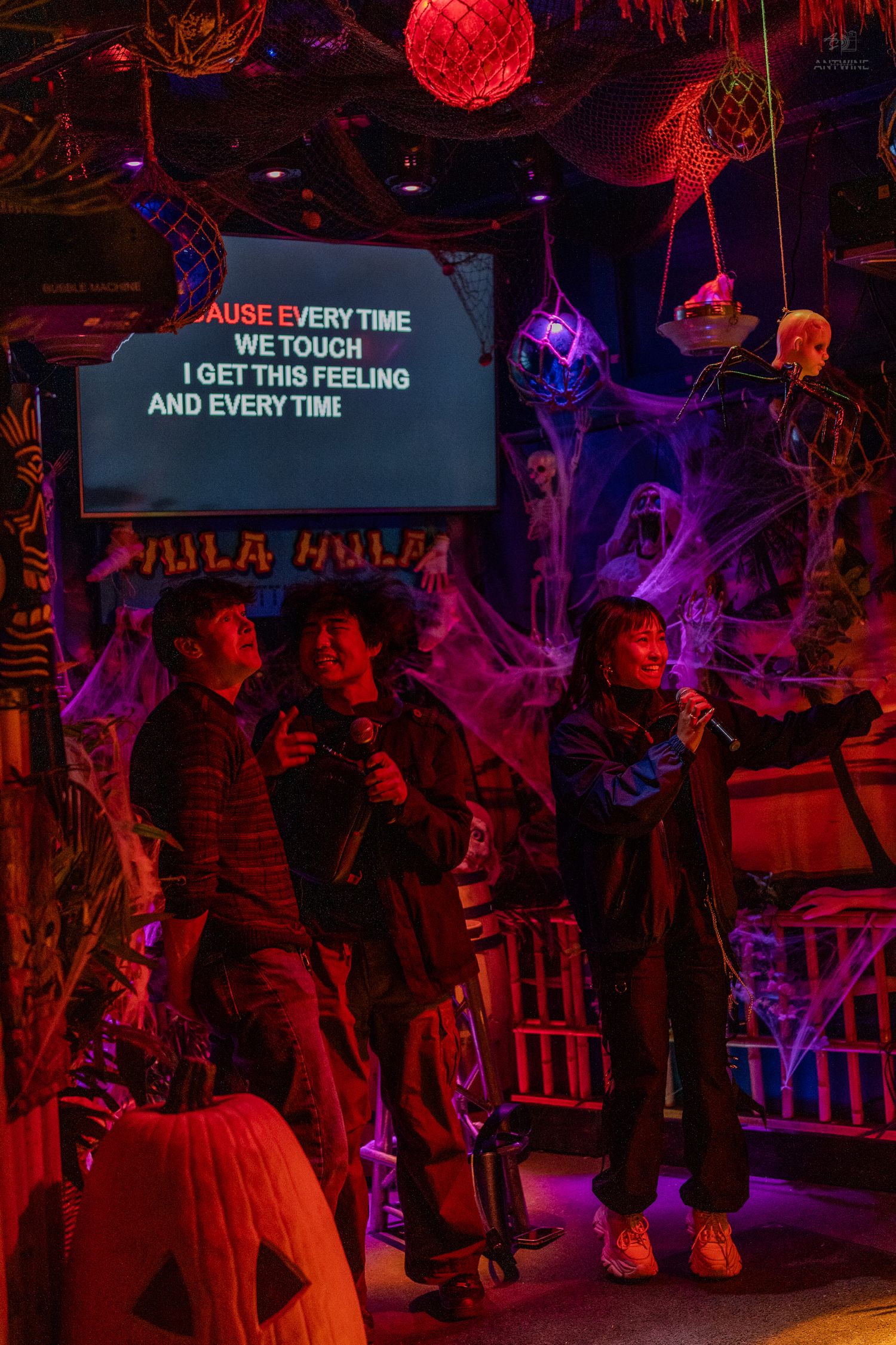 Group singing karaoke at Halloween-themed bar with spooky decorations, cobwebs, and colorful neon lights.