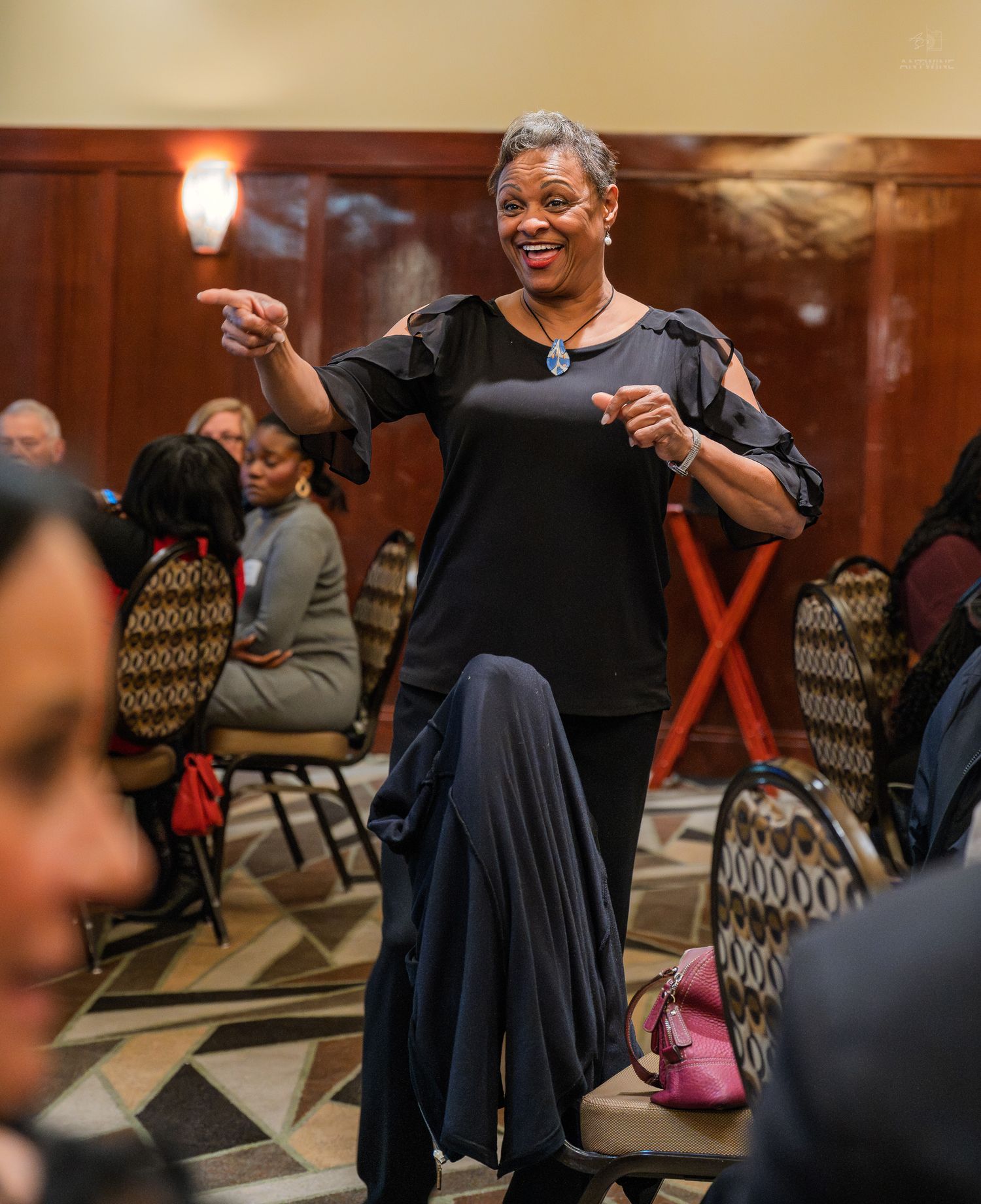 A woman in black attire smiles and points at a gathering in an elegant banquet hall.