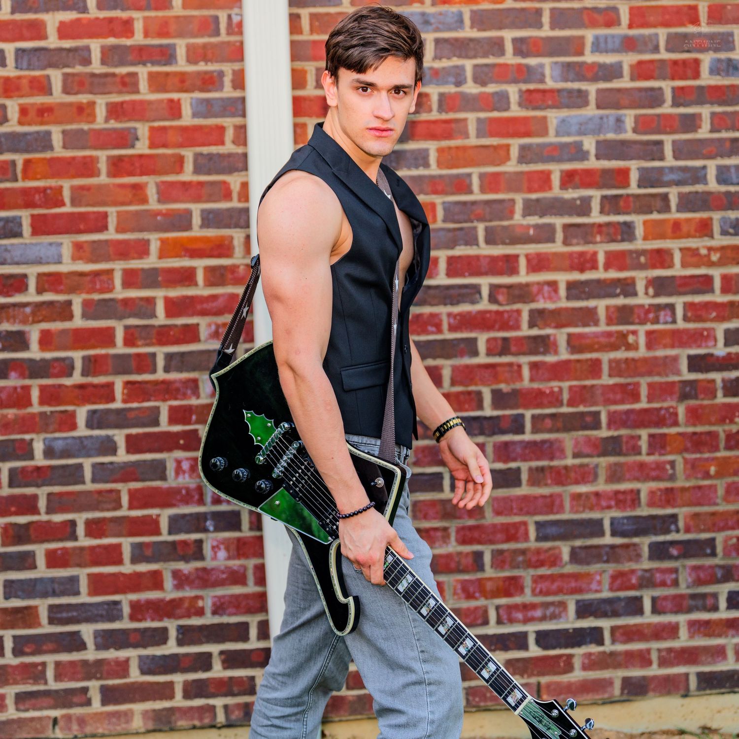 Young man in black vest holding an electric guitar, posing against a red brick wall outdoors.