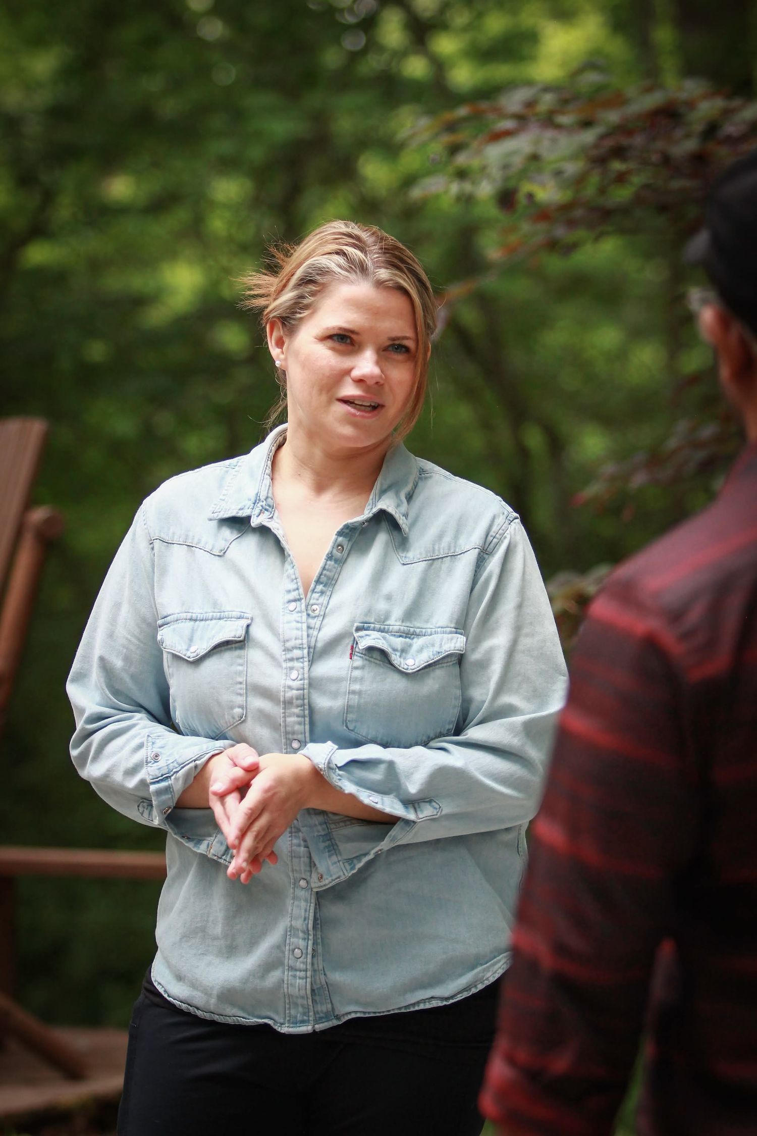 Blonde woman in denim shirt speaking outdoors in a lush green wooded background.