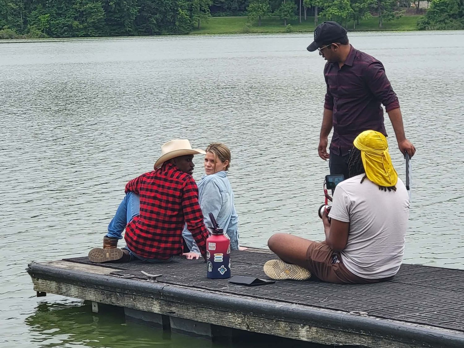 A diverse group of four people sit and stand on a wooden dock by a calm green lake on an overcast day.