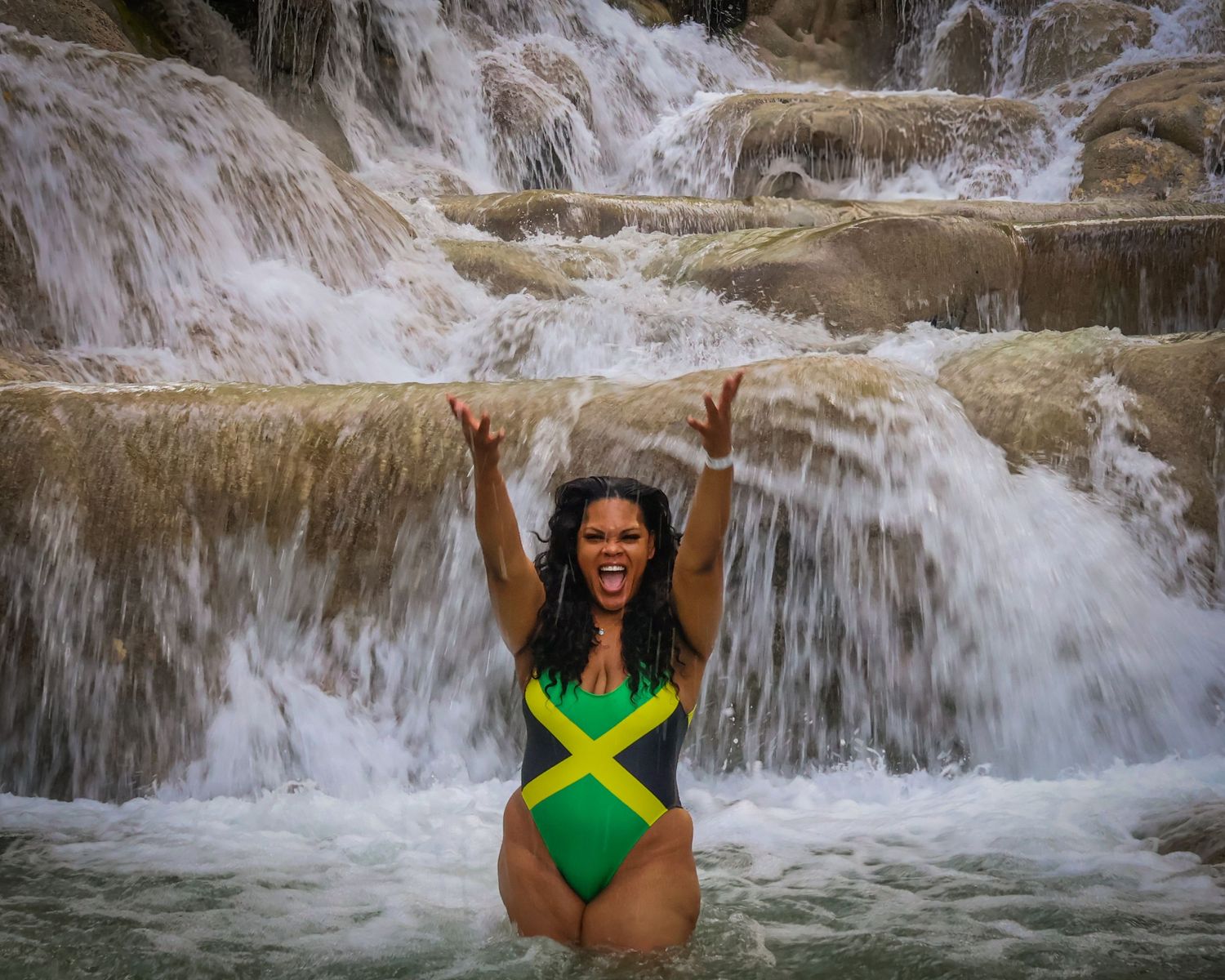 A female standing infront of a rushing waterfall throwing water up into the air.
