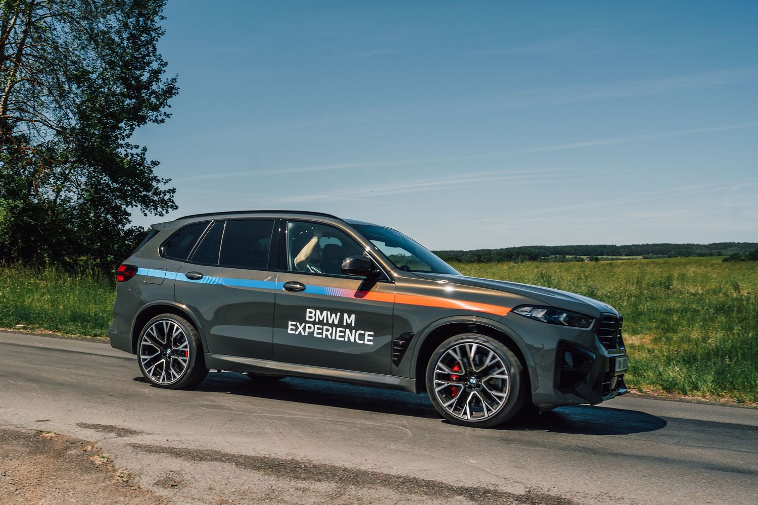 BMW X5 M Competition with BMW M Experience wrap parked on a sunny rural road near green fields.