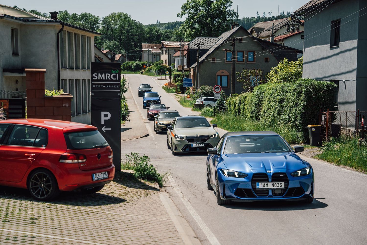 Blue BMW M2 leads a convoy of cars through a small European village street on a sunny day.