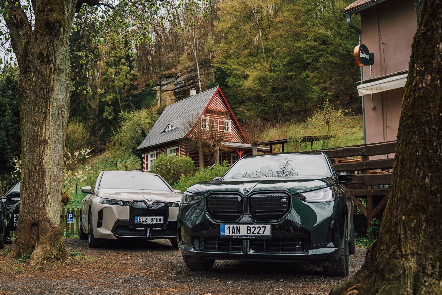 Two BMW electric SUVs parked on a gravel driveway surrounded by autumn trees near a charming cottage.