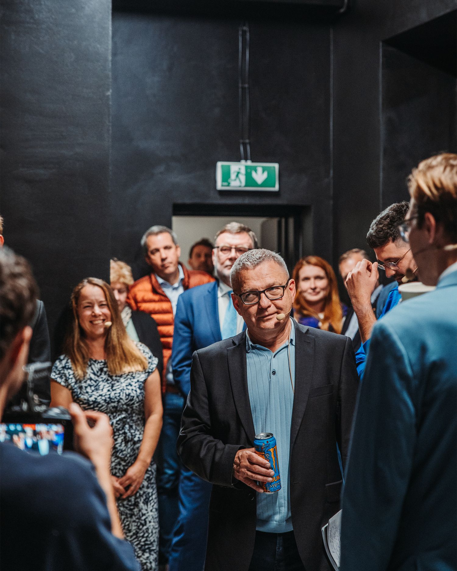 Older man in dark blazer holding a drink can while mingling with a crowd at an indoor event.