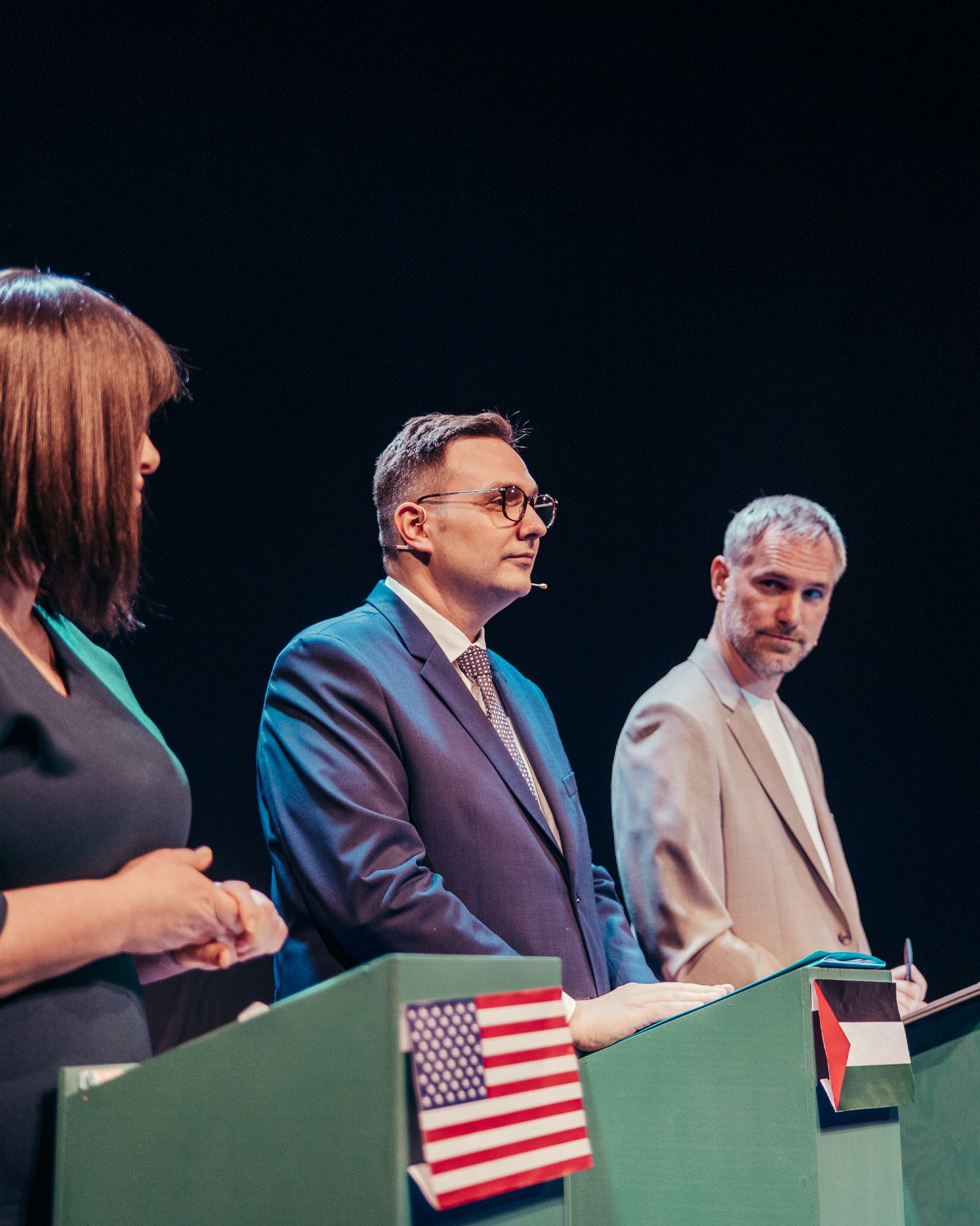 Two men and a woman standing at podiums with US and Palestinian flags during a formal debate.
