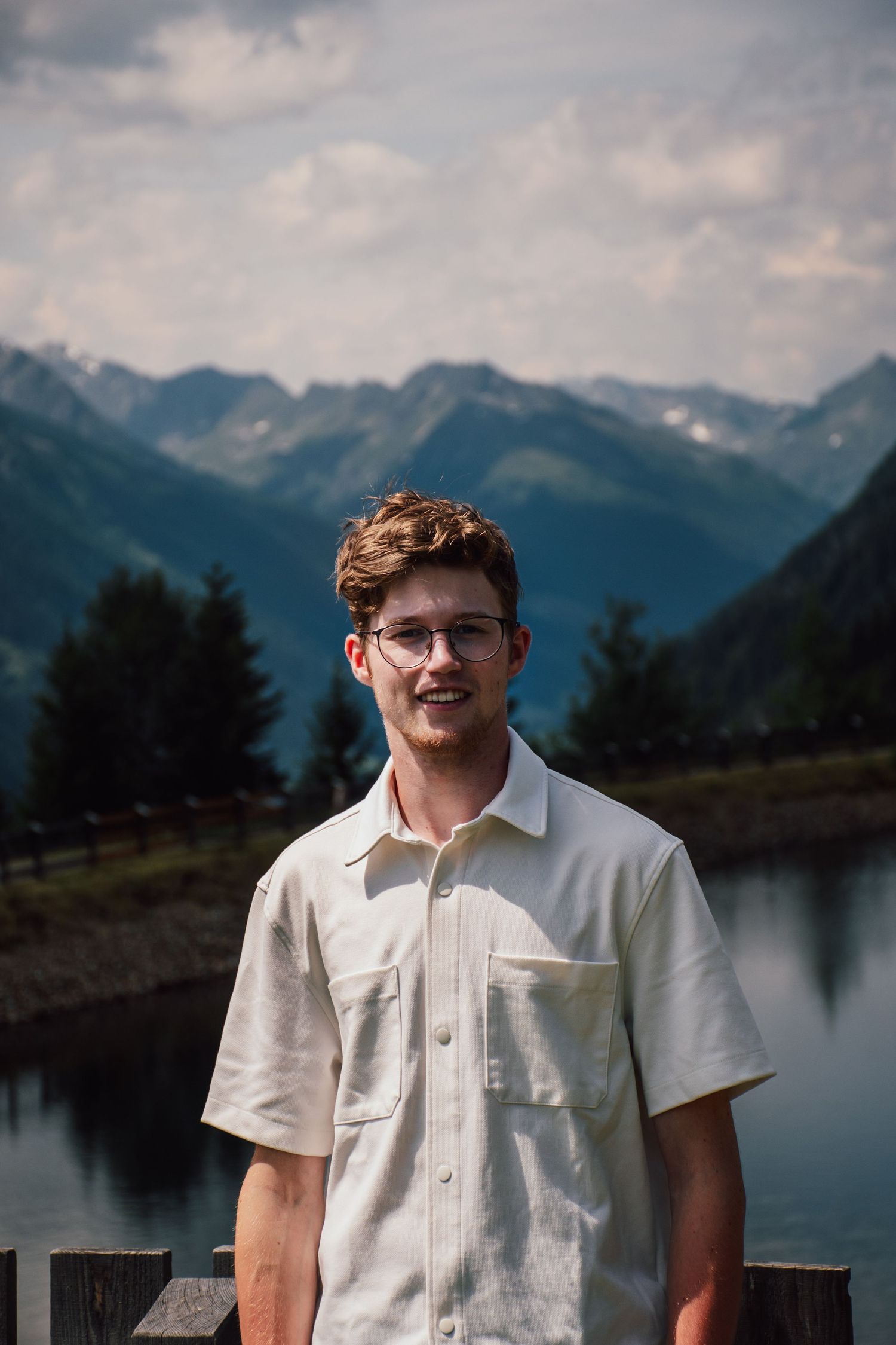 Young man with glasses in white shirt smiling before stunning mountain lake landscape.