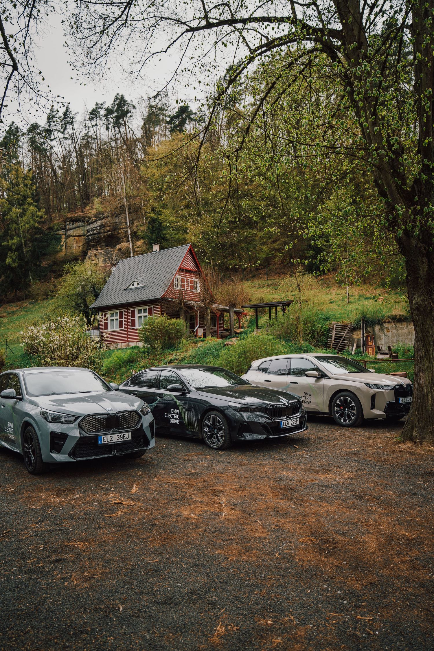 Three BMW electric vehicles parked side by side on gravel with a red cottage and green hillside backdrop.