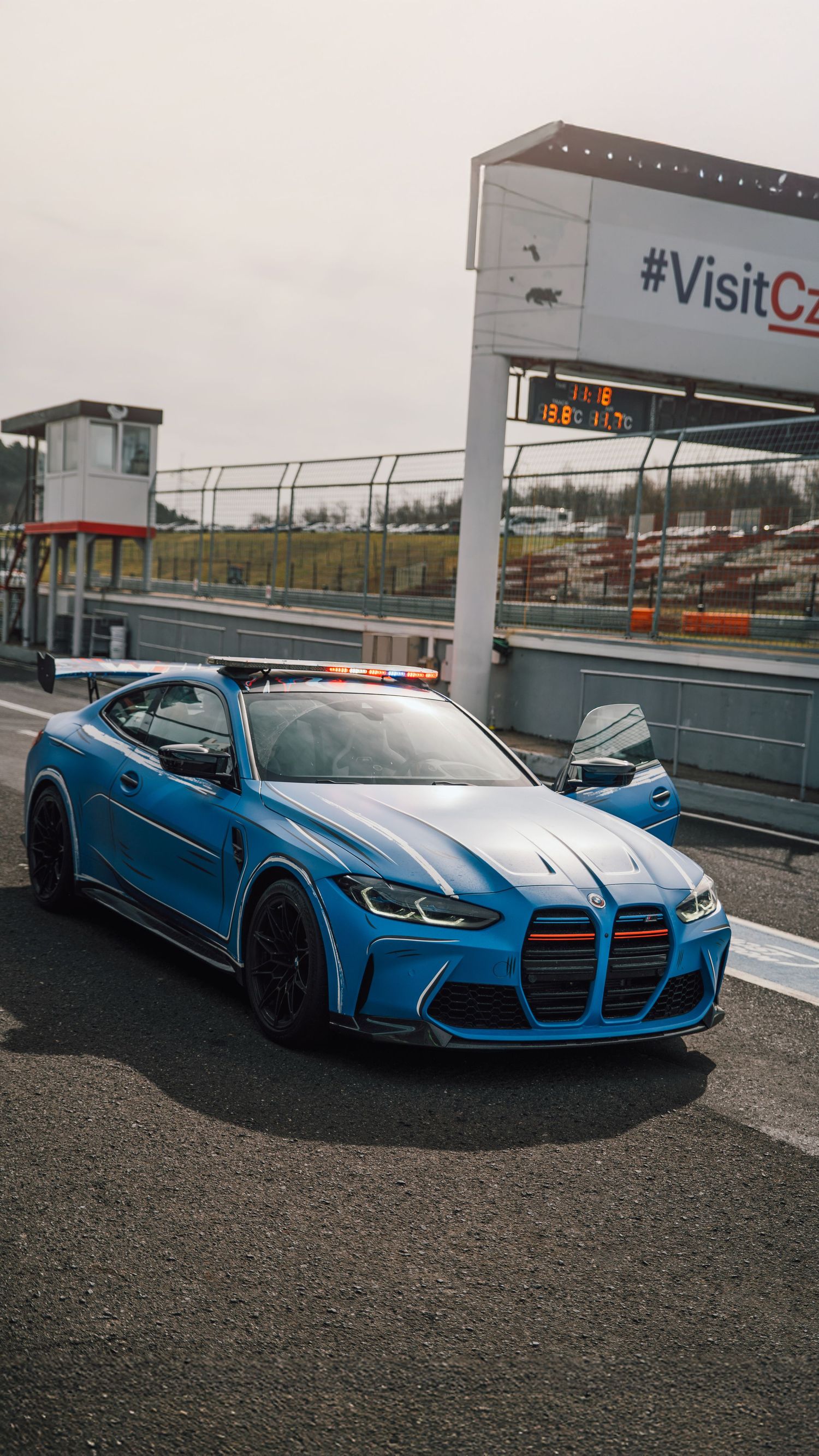 Blue BMW M4 GT safety car with racing spoiler and light bar parked in pit lane at a racing circuit.