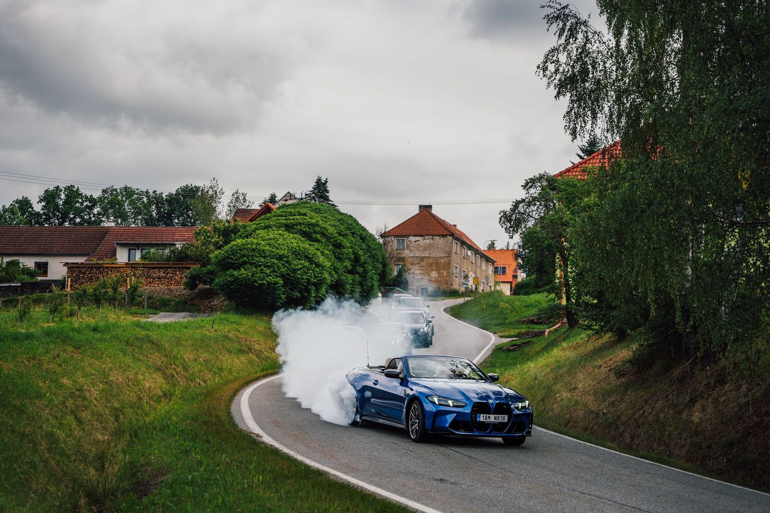Blue BMW M4 performing a dramatic tire burnout on a curved village road under an overcast sky.
