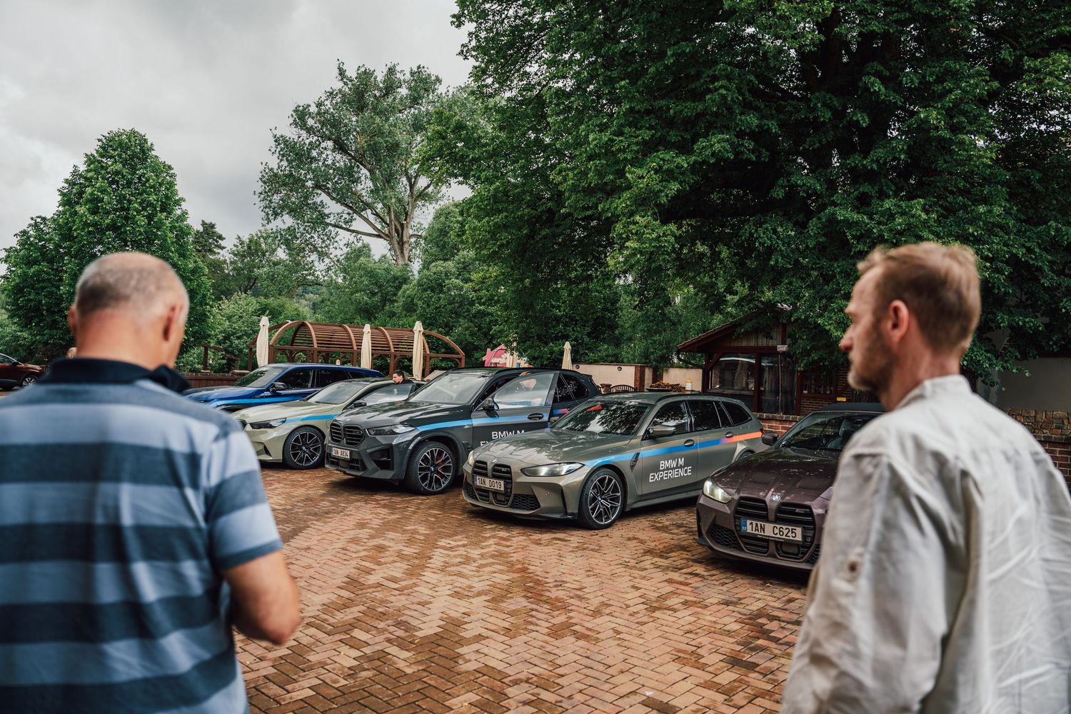 Group of people viewing multiple BMW M Experience vehicles parked on brick lot under cloudy sky.