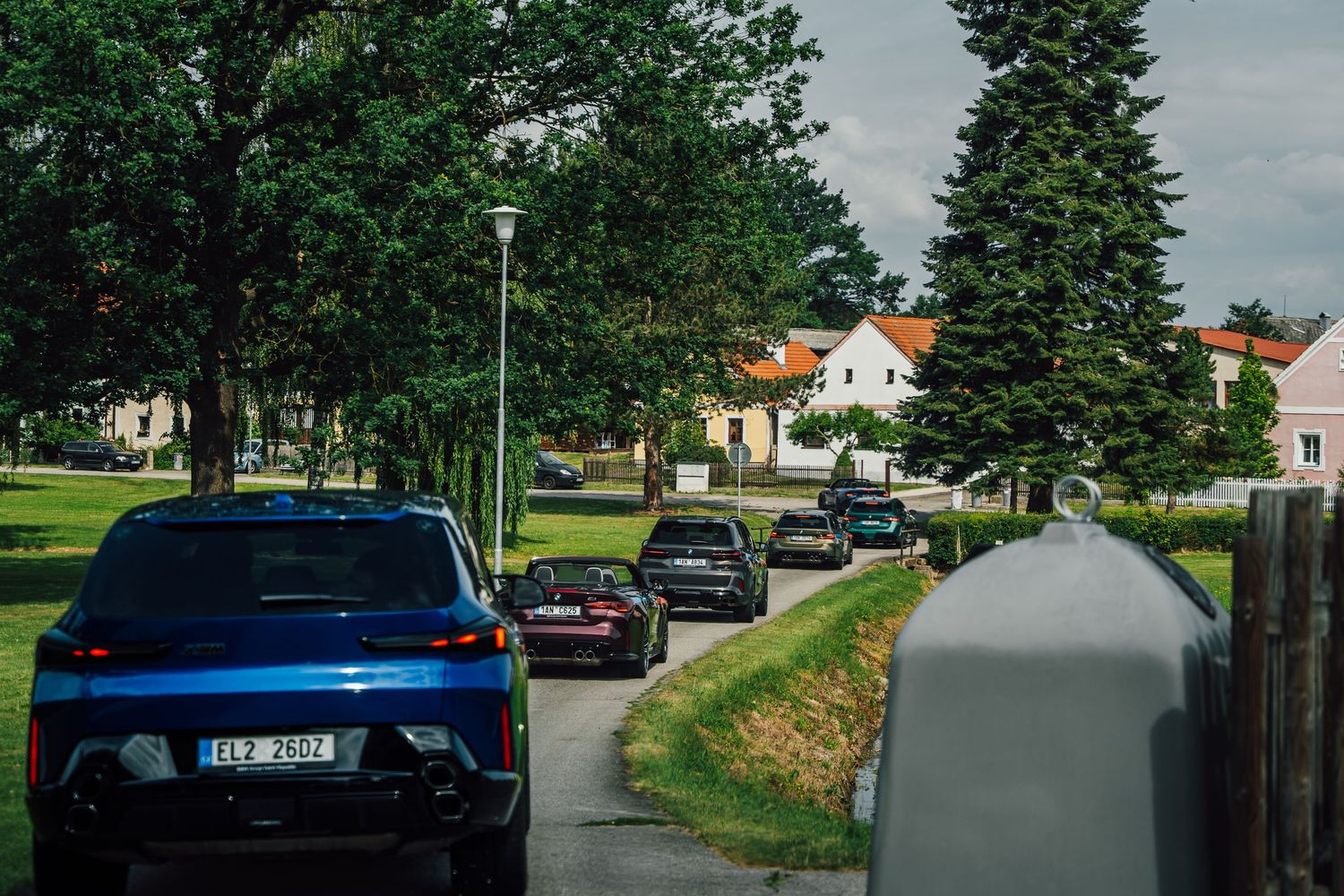 A convoy of BMW M cars driving through a tree-lined residential street in a European village.