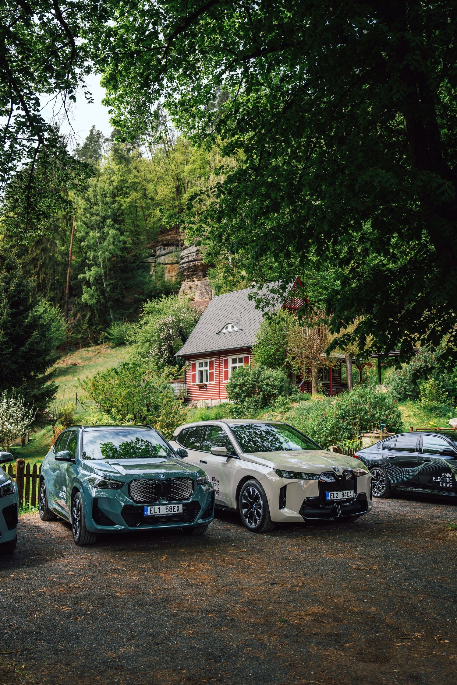 Two BMW electric SUVs parked on a gravel lot with a red stone cottage nestled in a forested hillside.