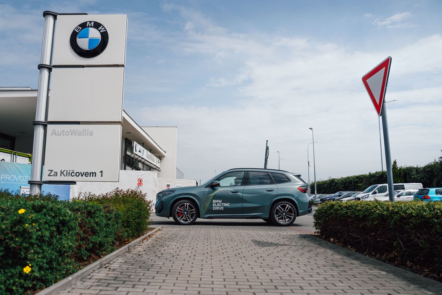 Dark green BMW iX5 parked outside a BMW dealership beside a yield sign on a sunny day.