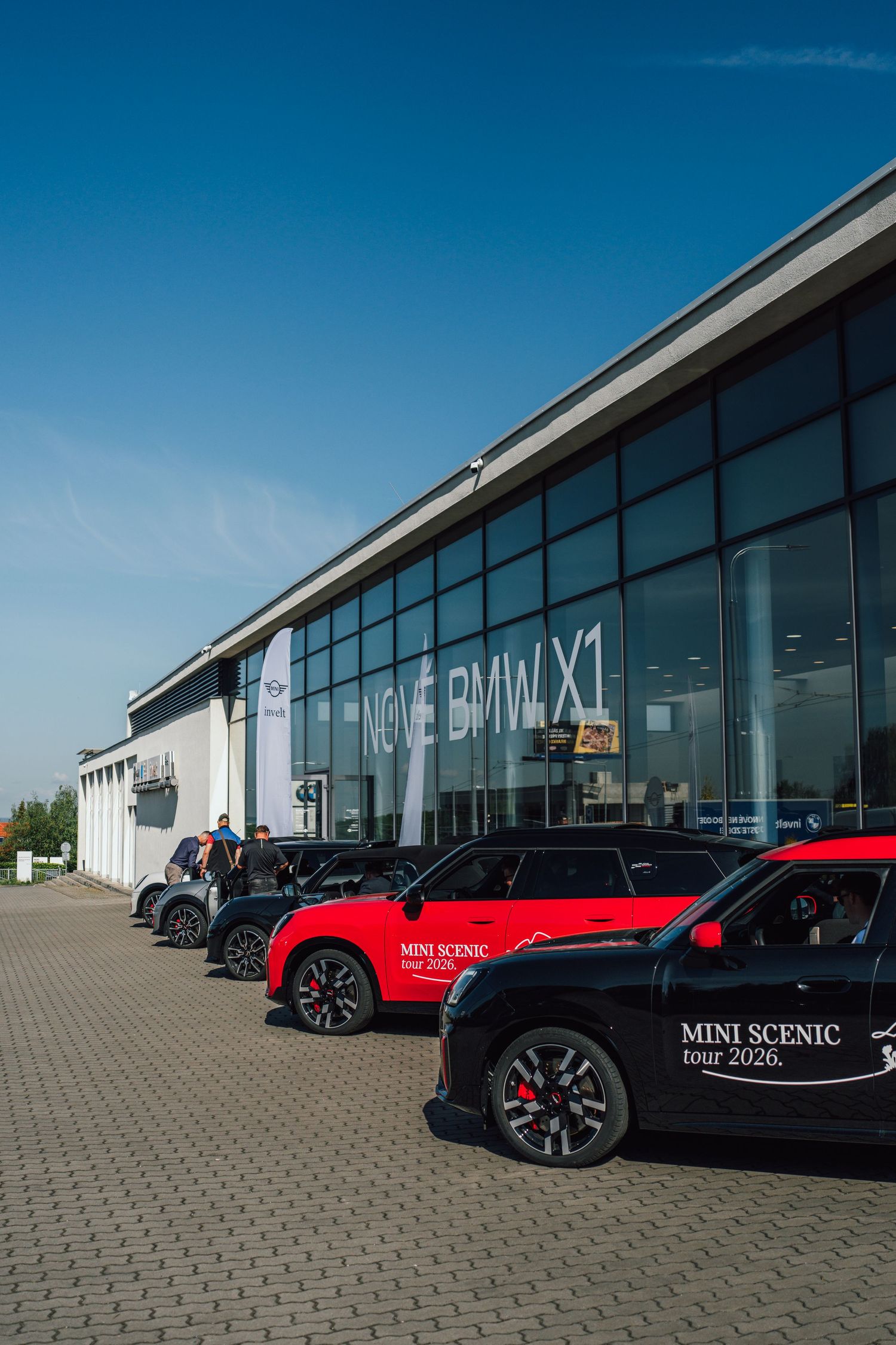 MINI Scenic tour 2026 branded cars lined up outside BMW X1 dealership under blue sky.