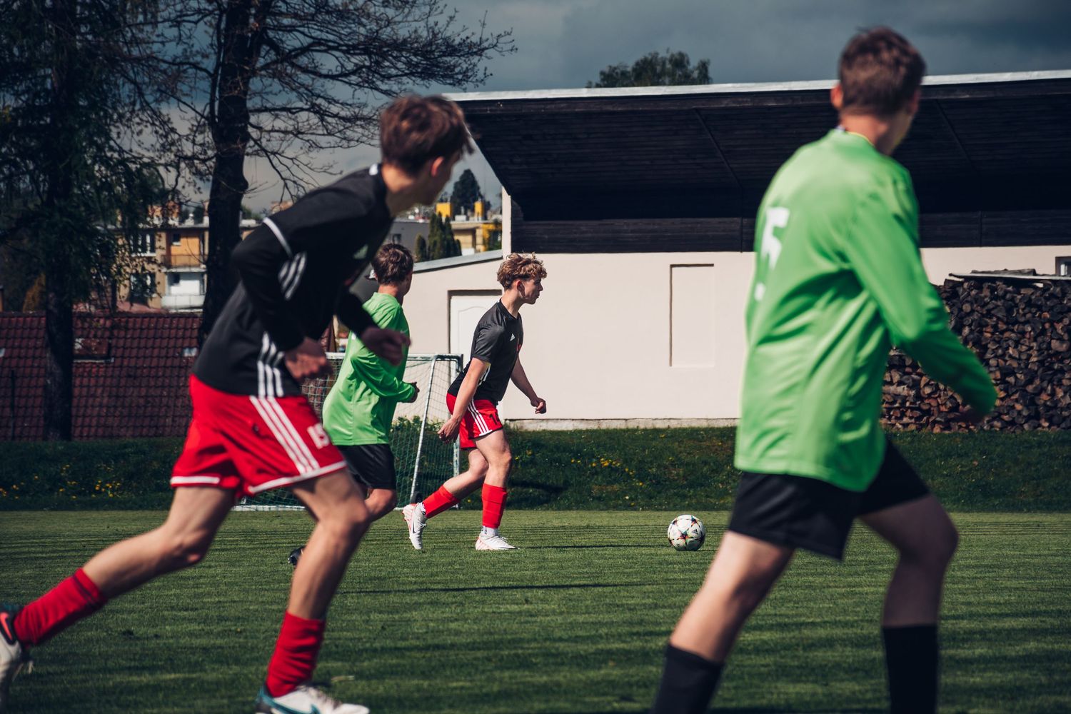 Youth soccer players in black, red, and green uniforms compete for the ball on a sunny green field.
