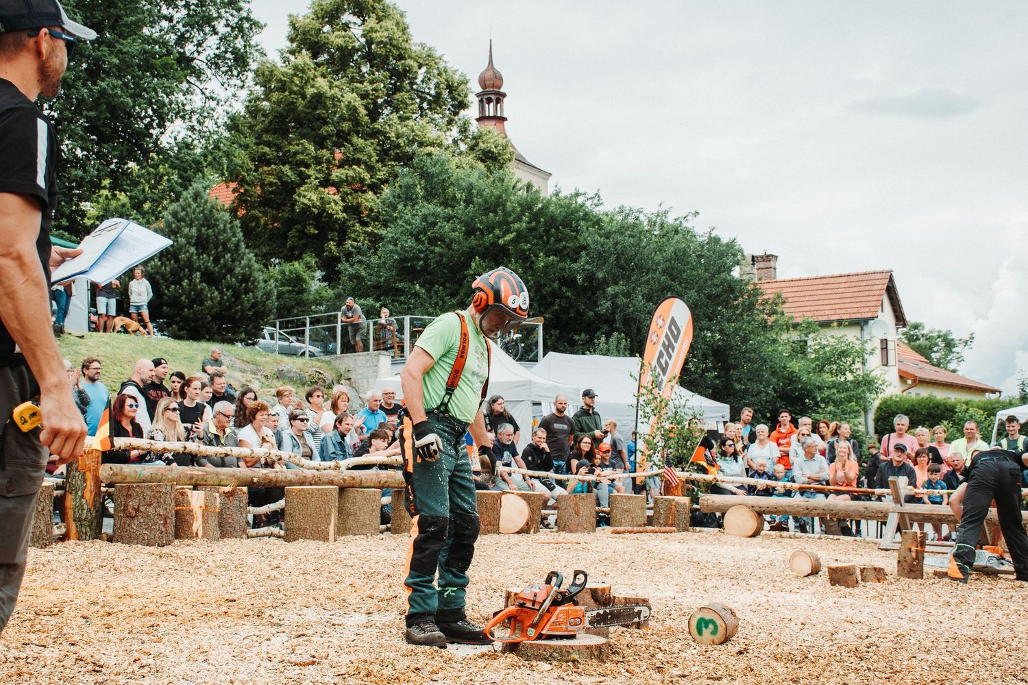 Lumberjack competitor stands with chainsaw before large crowd at outdoor timber sports competition.