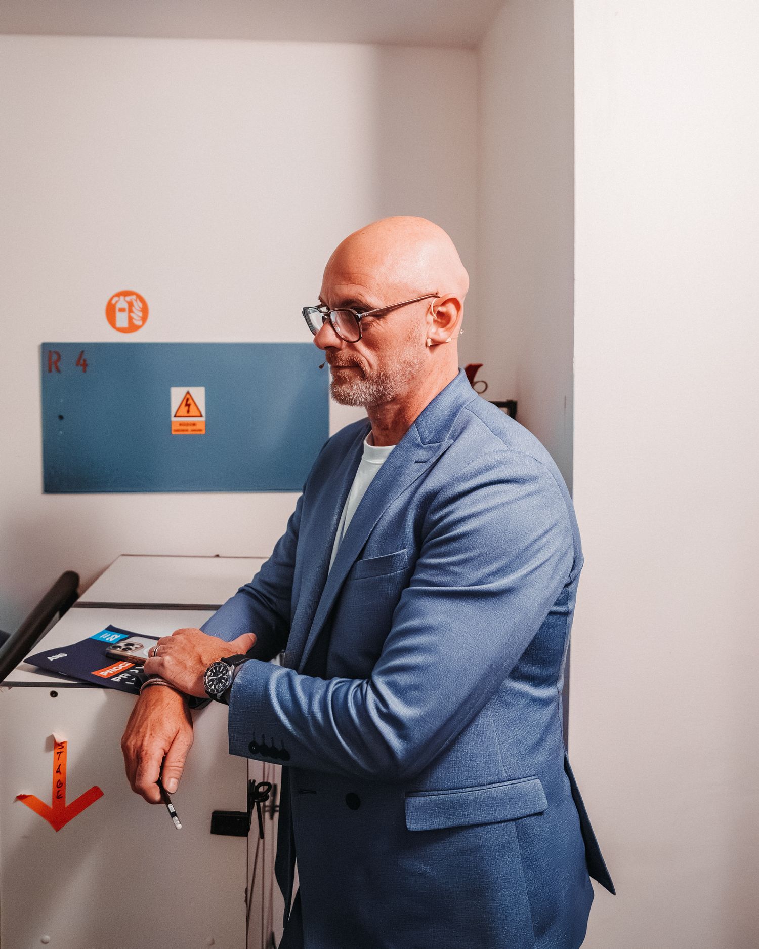 Bald man with glasses in a blue blazer leaning on equipment in a white industrial room.