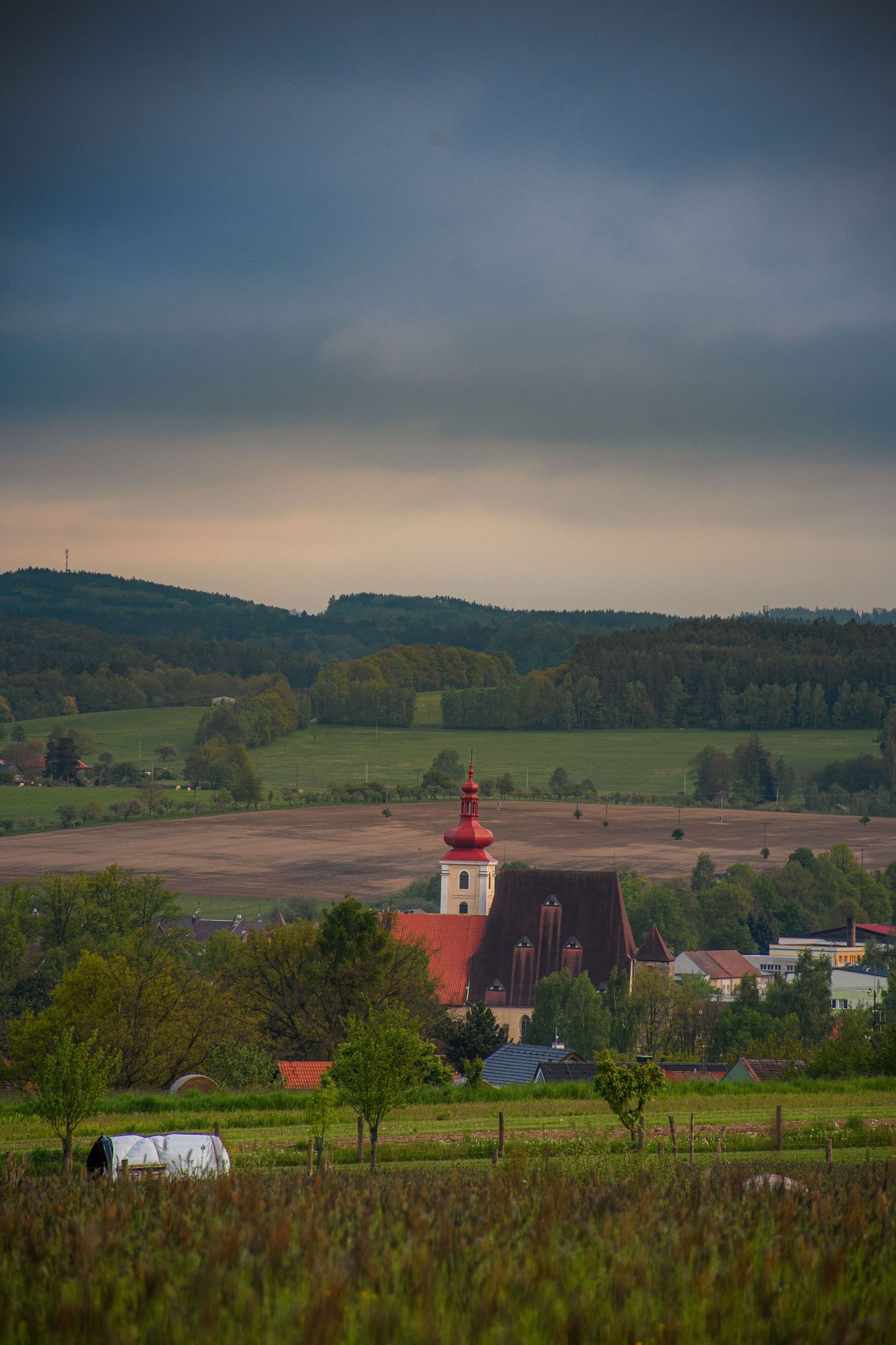 A quaint European village church with a red roof sits amid rolling green fields under a stormy sky.