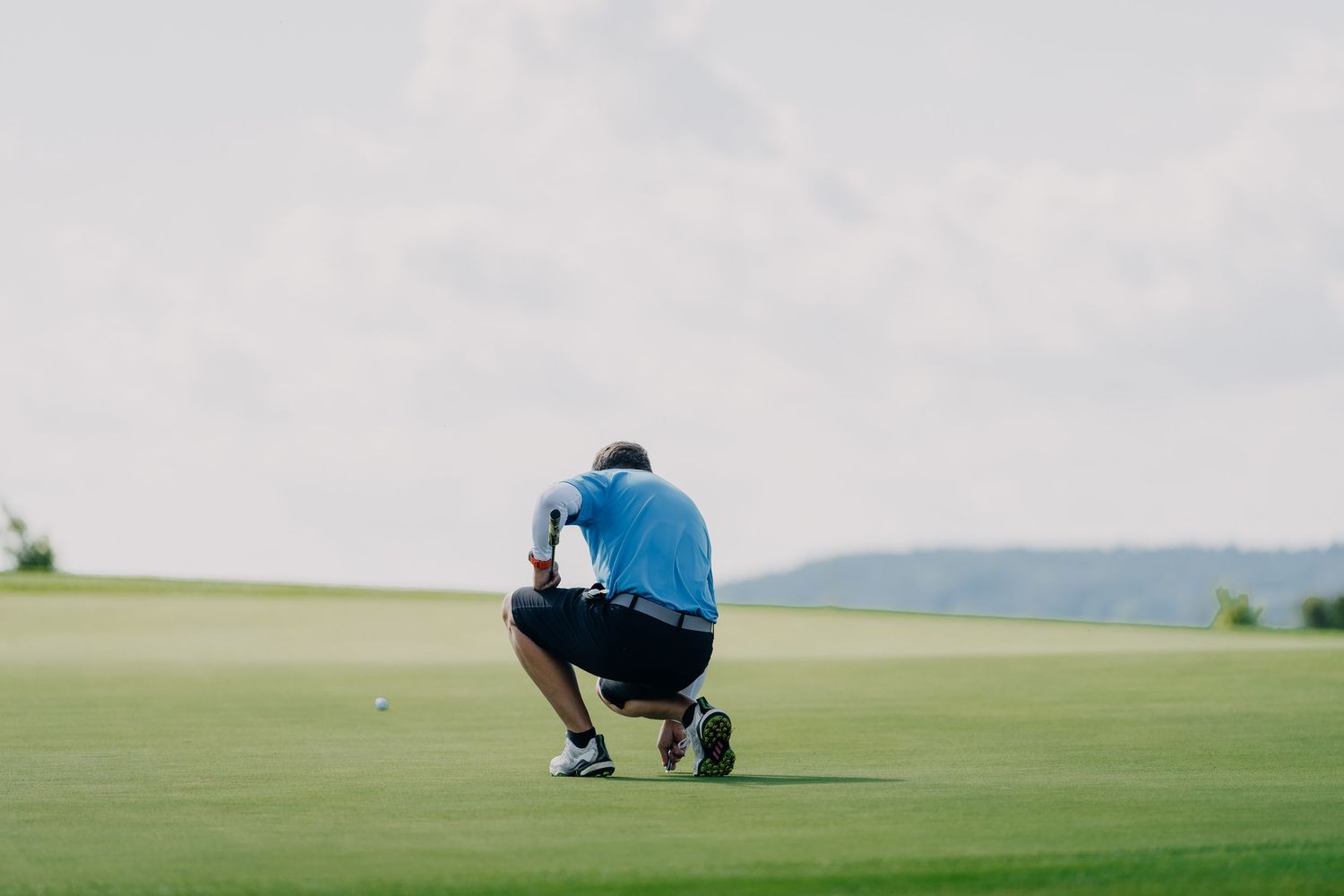 Golfer crouching on green grass putting green, analyzing shot line on a golf course under cloudy sky.