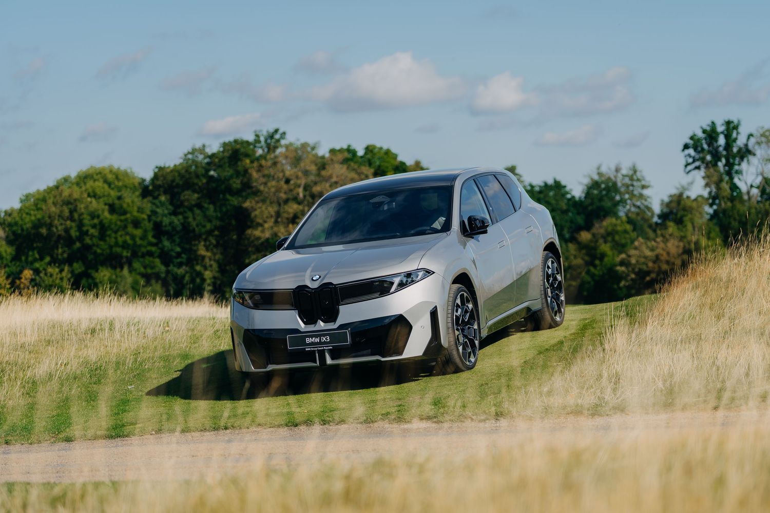 A silver BMW X3 SUV driving on a rural road surrounded by lush green trees and golden grasslands under a blue sky.