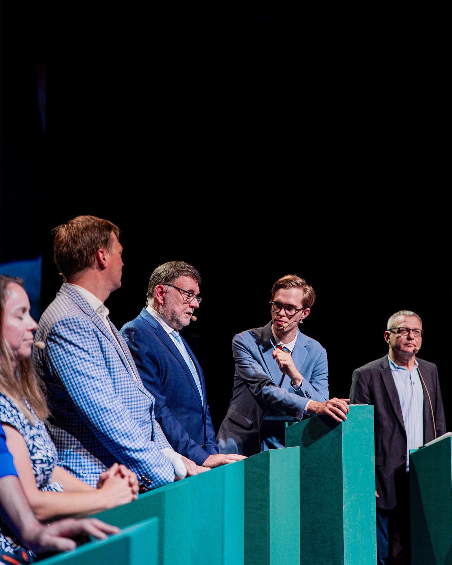 Five panelists stand at green podiums on a dark stage during a formal debate.