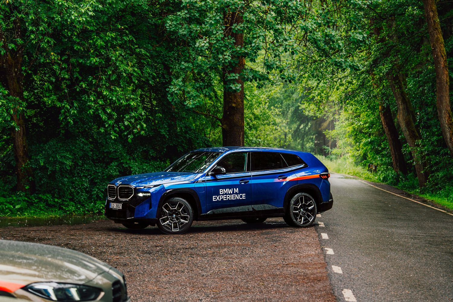 Blue BMW XM SUV parked on a tree-lined road with lush green forest backdrop.