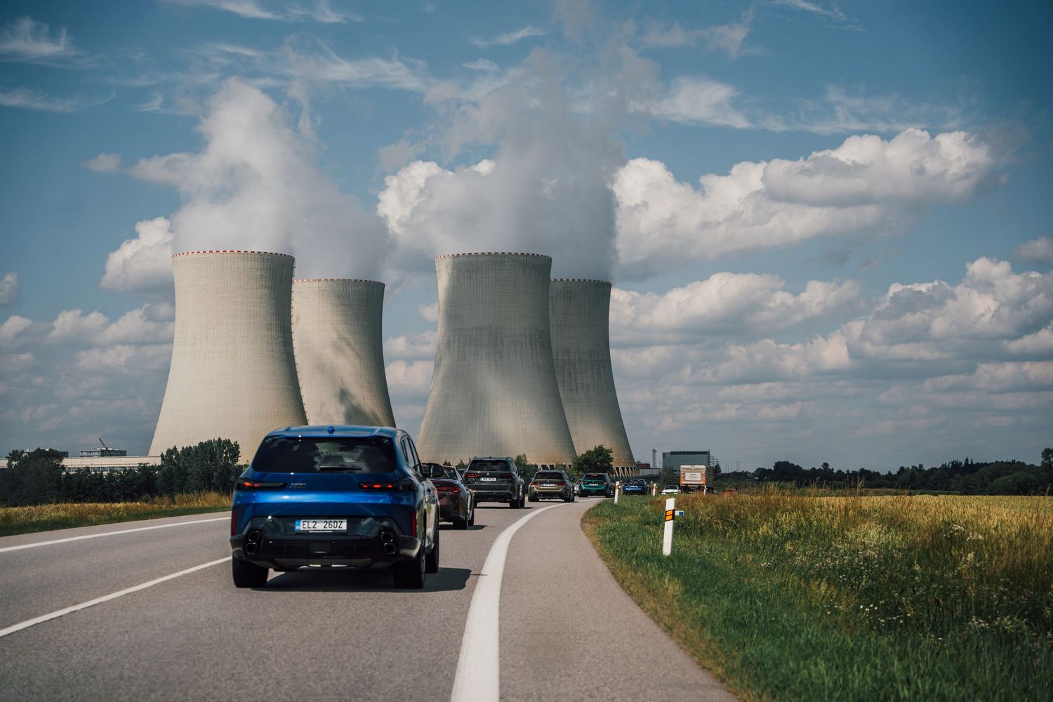 Cars driving on a highway past large nuclear power plant cooling towers emitting steam under a cloudy sky.