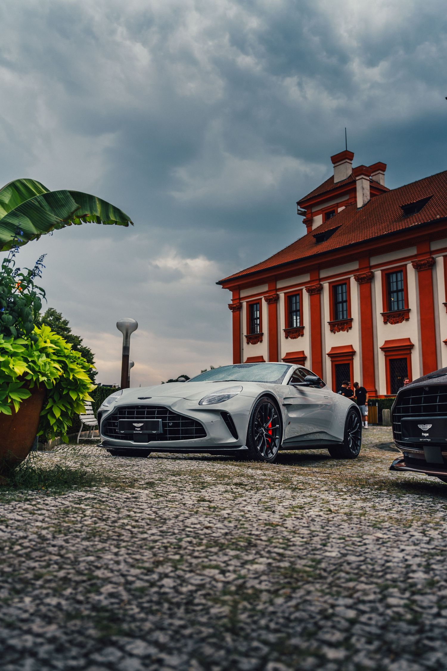 Silver Aston Martin Vantage roadster parked on cobblestones in front of a red Baroque-style manor under stormy skies.