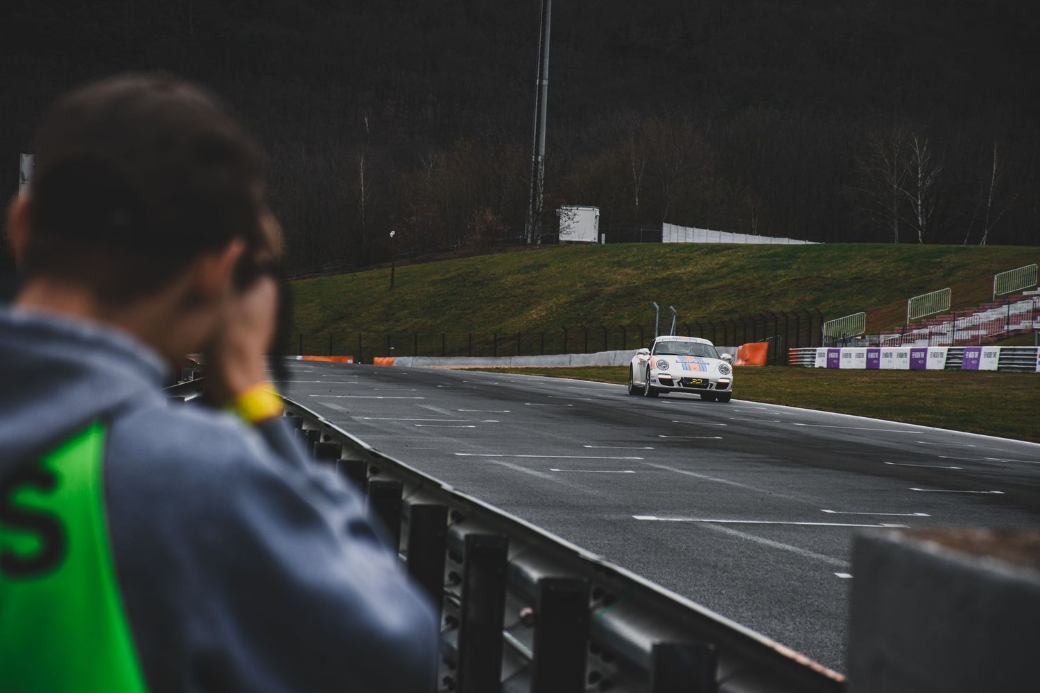 Spectator watches a white Porsche race car speed along a wet racing circuit.