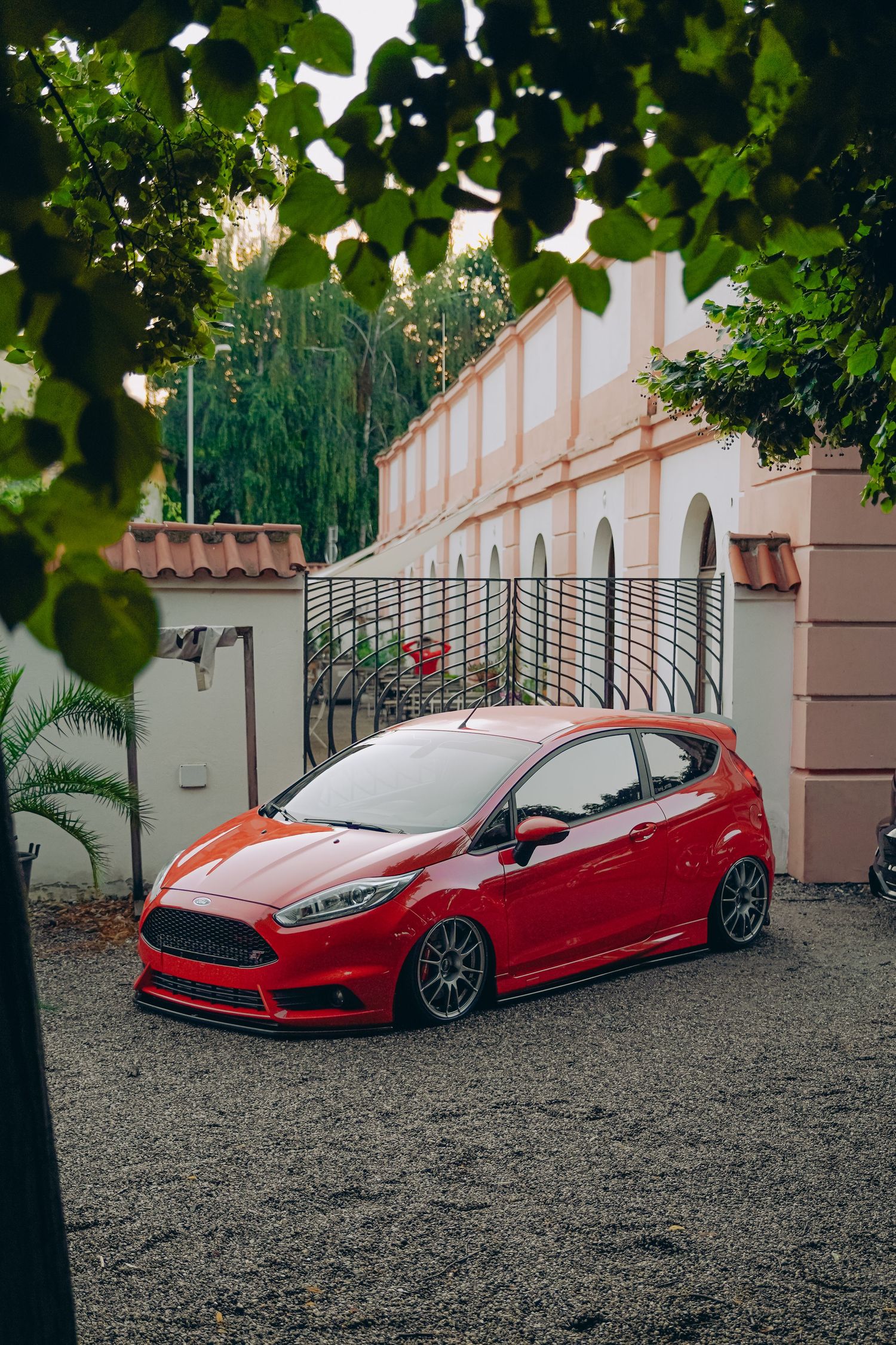 Red Ford Fiesta ST lowered on gray alloy wheels parked on gravel outside Mediterranean-style villa.