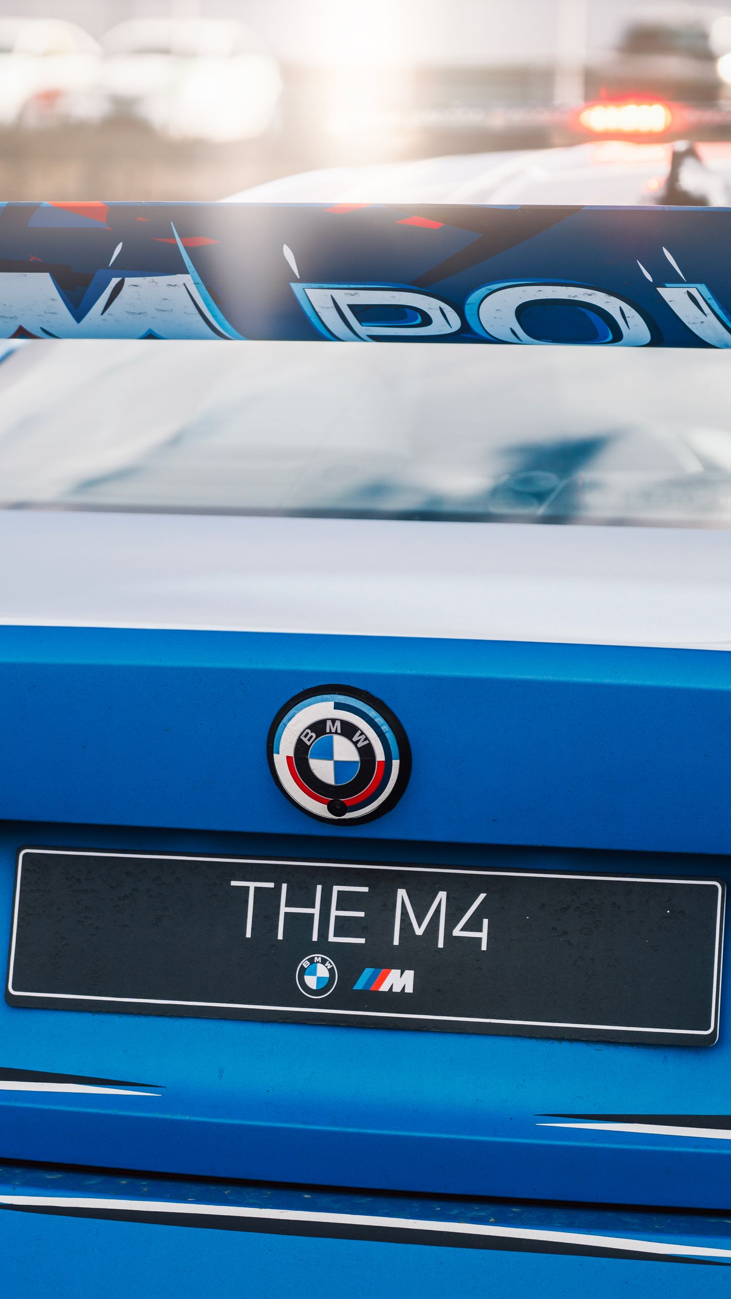 Rear view of a blue BMW M4 showing the iconic roundel badge and THE M4 license plate in sunlight.