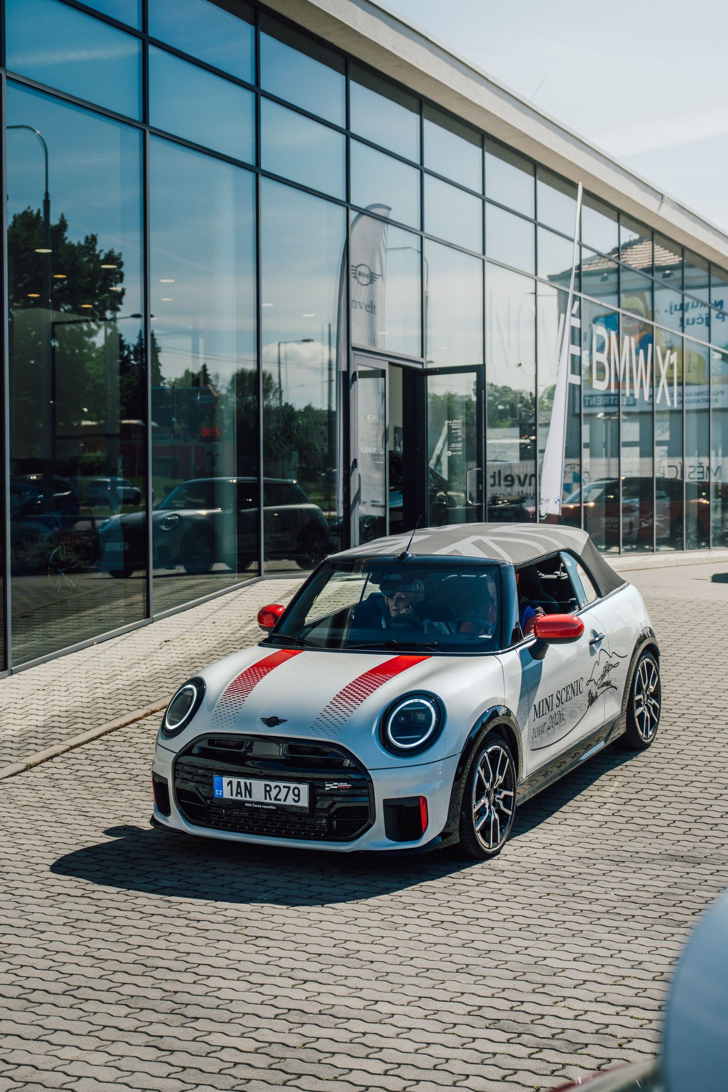 White MINI John Cooper Works convertible with red stripes parked outside a BMW dealership on a sunny day.