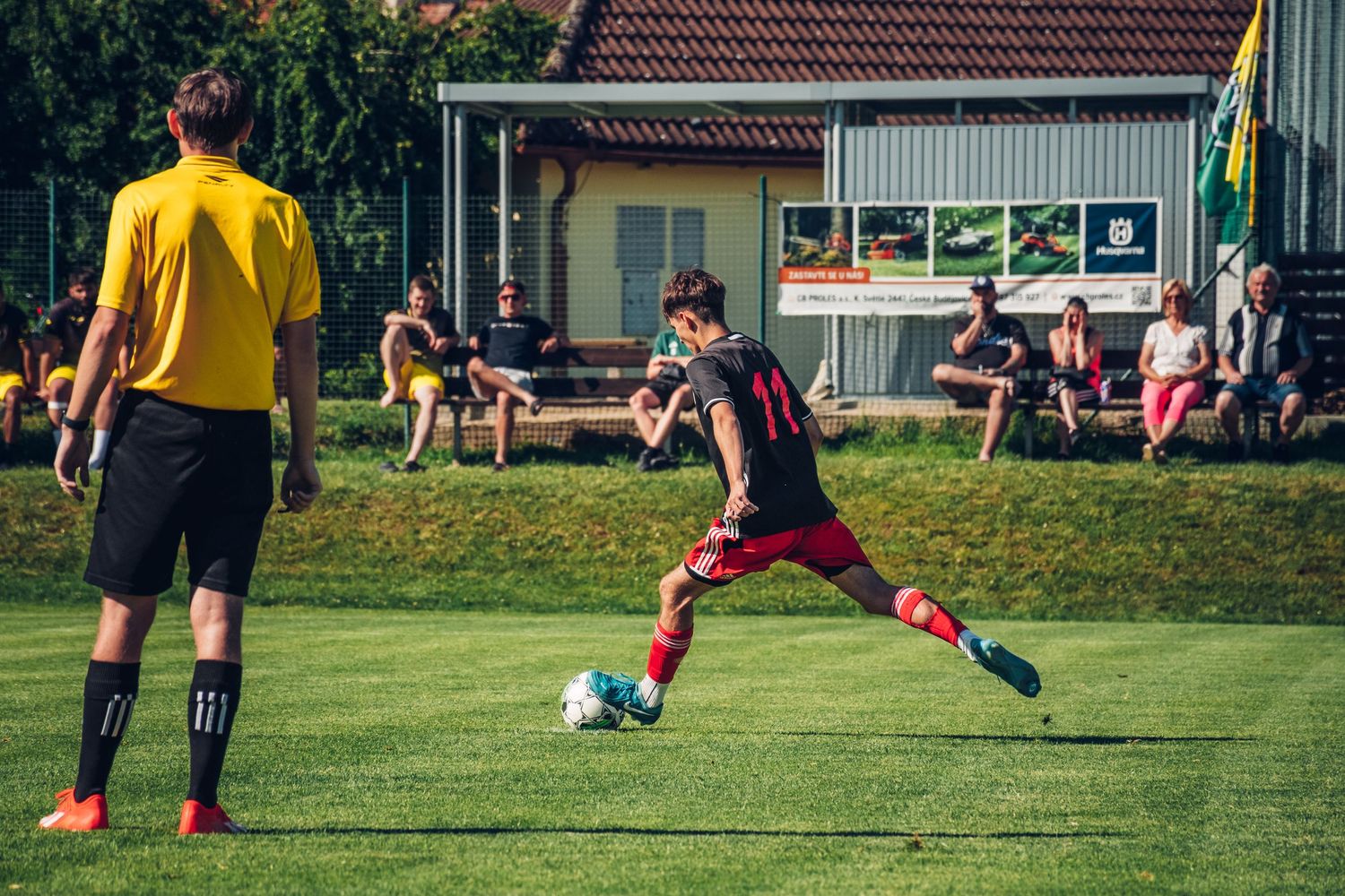 Young soccer player in red and black uniform prepares to kick ball near sideline referee.
