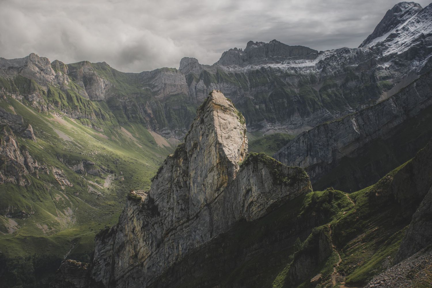 A dramatic solitary rock spire rises between green mountain valleys under a dark moody cloudy sky.