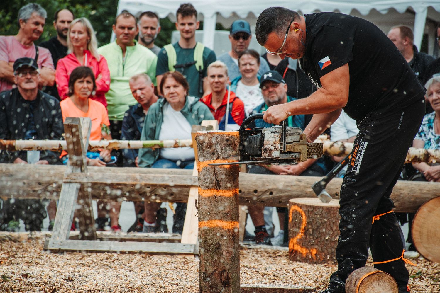 Competitor uses chainsaw on multiple logs while crowd watches at outdoor lumberjack sports event.