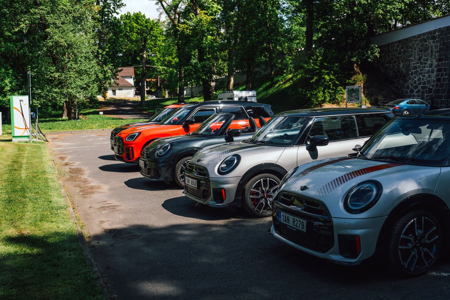 Row of colorful MINI Cooper cars parked in a shaded lot surrounded by lush green trees.