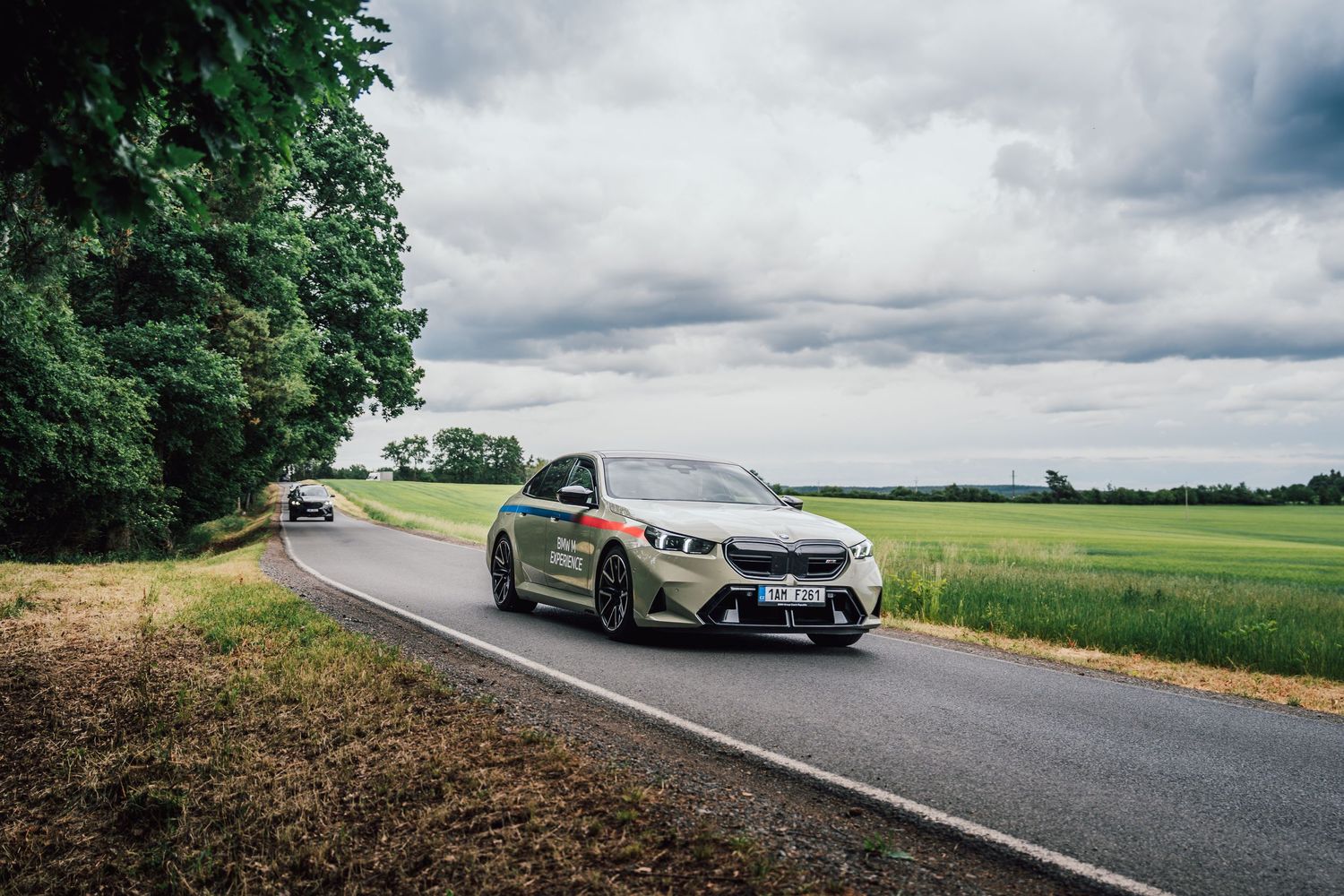 BMW 5 Series in military green driving on a rural road flanked by green fields.