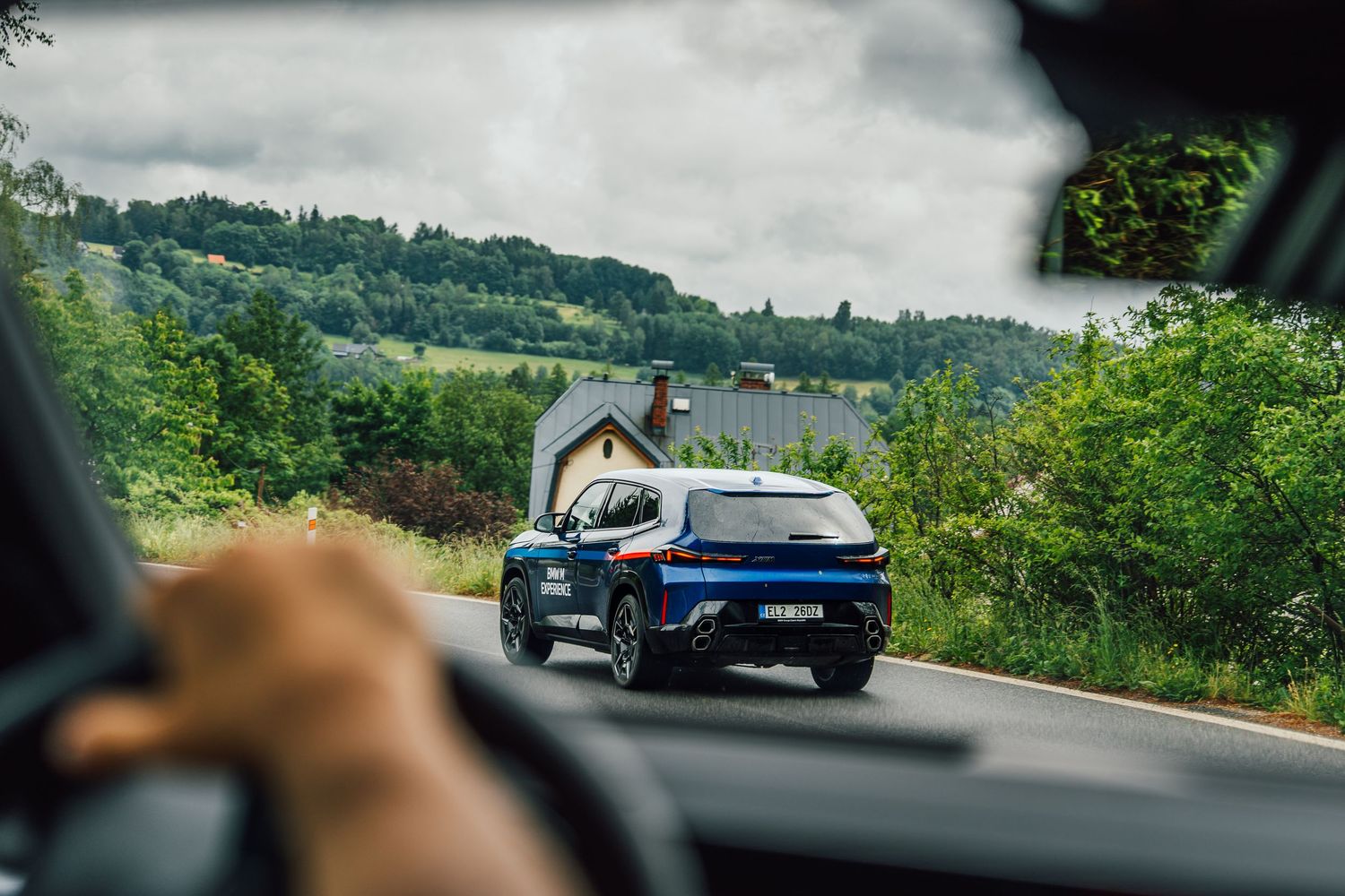 Blue BMW M Experience SUV driving on a rural road through scenic green hills and farmland.