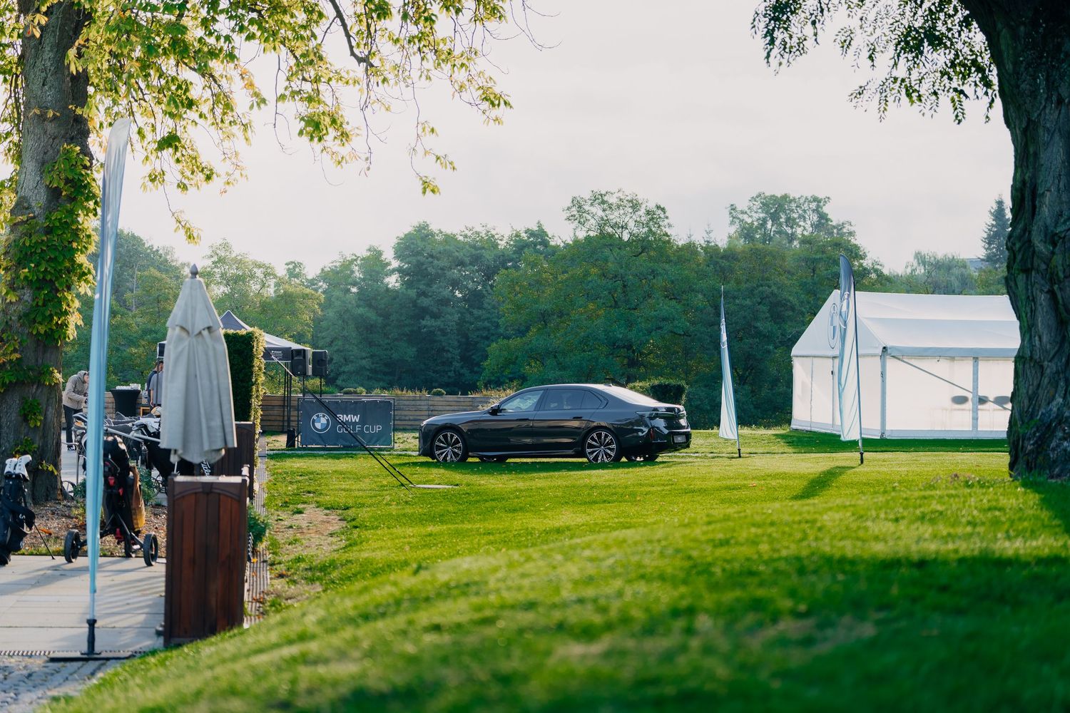 Black luxury car parked on lush green lawn near white event tent and trees at an outdoor gathering.