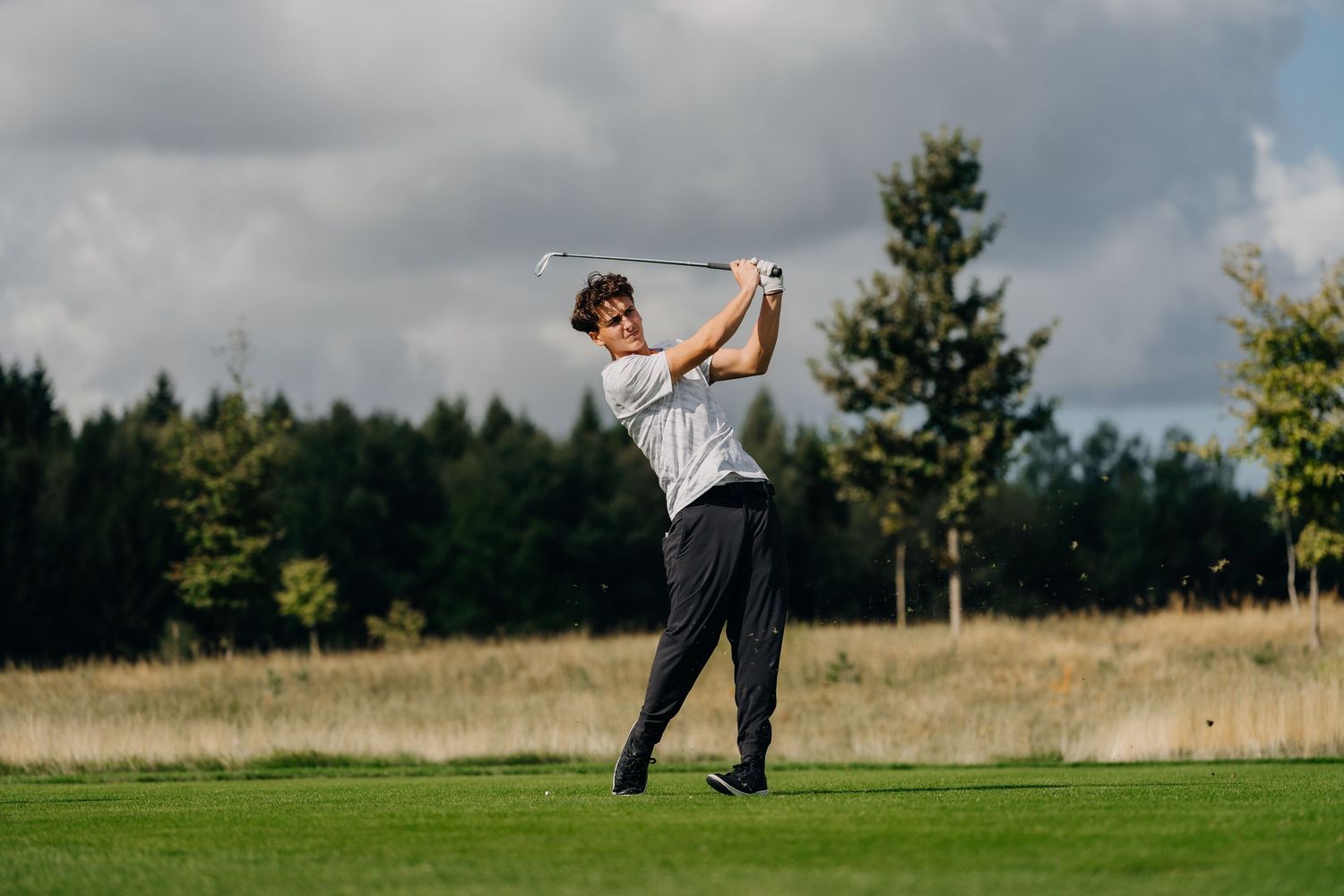 Young golfer swings a club on a lush golf course with trees and cloudy sky in the background.